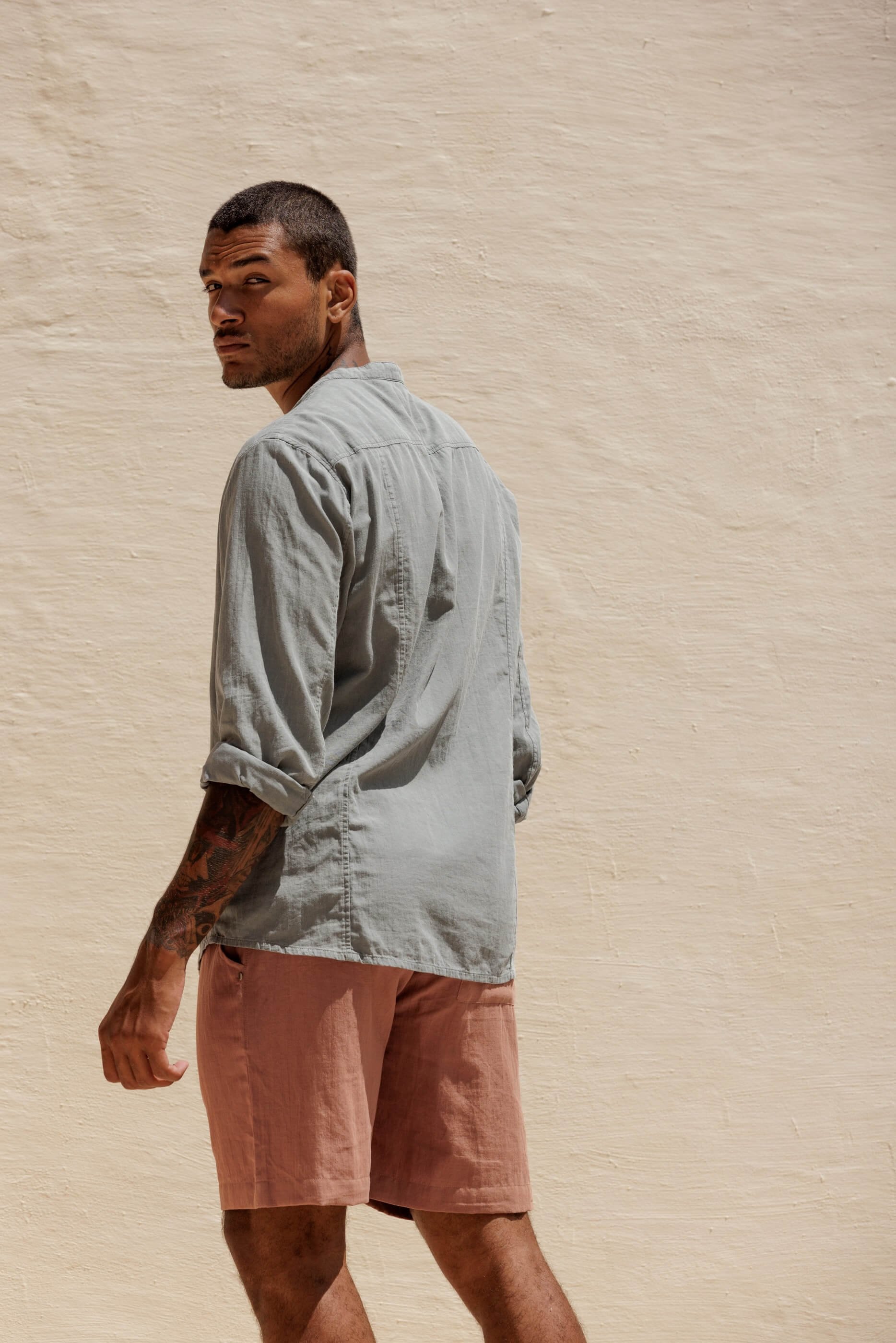 Three-quarter rear view from the knee up of the model wearing the terracotta Renclì shorts paired with the Jordi extra-fine cotton shirt in Sage Stone grey-green.