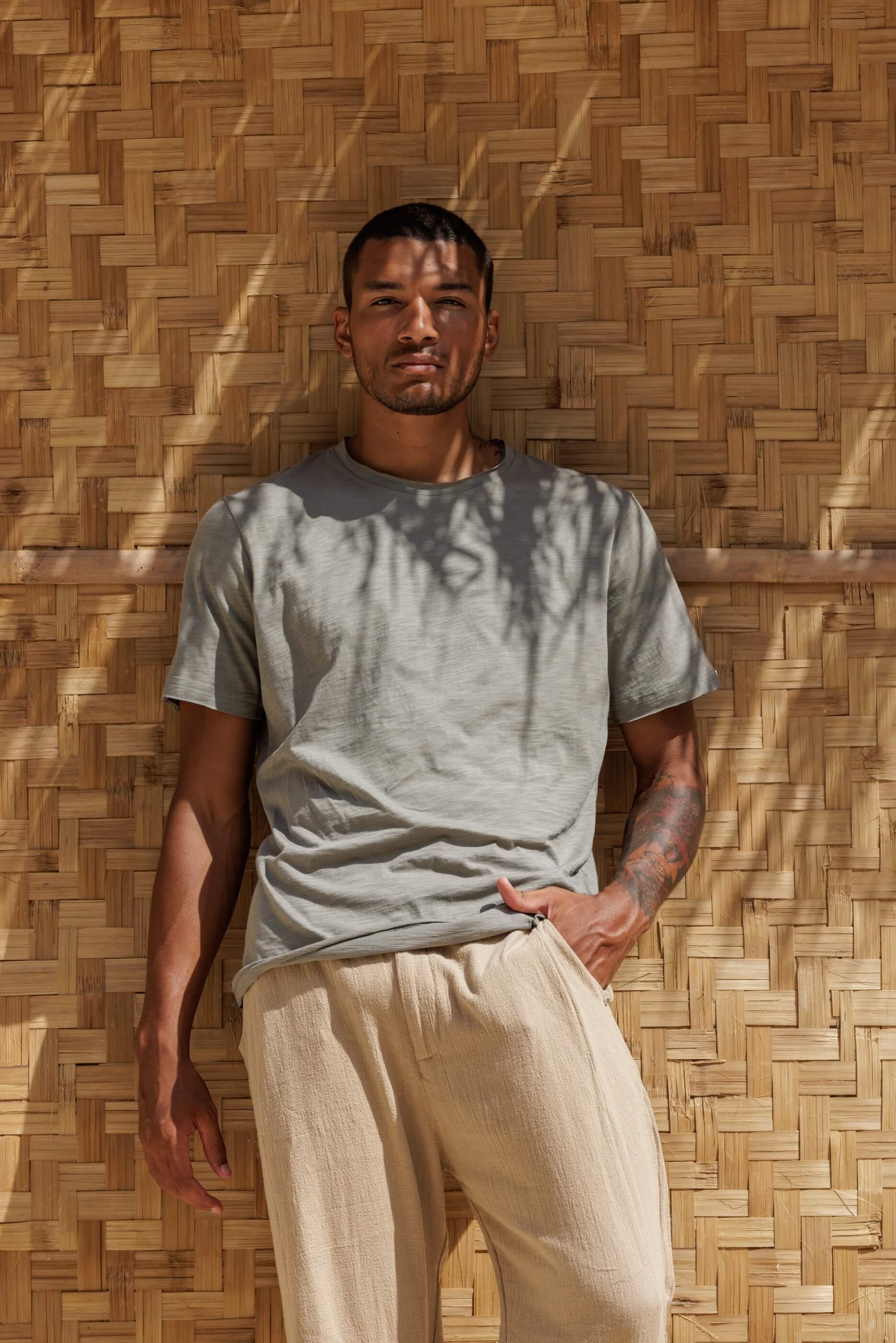 Medium shot of a model wearing the Nestor T-shirt in Dust Grey green, with artistic plant shadows showcasing the contemporary loose fit and raw-cut collar.