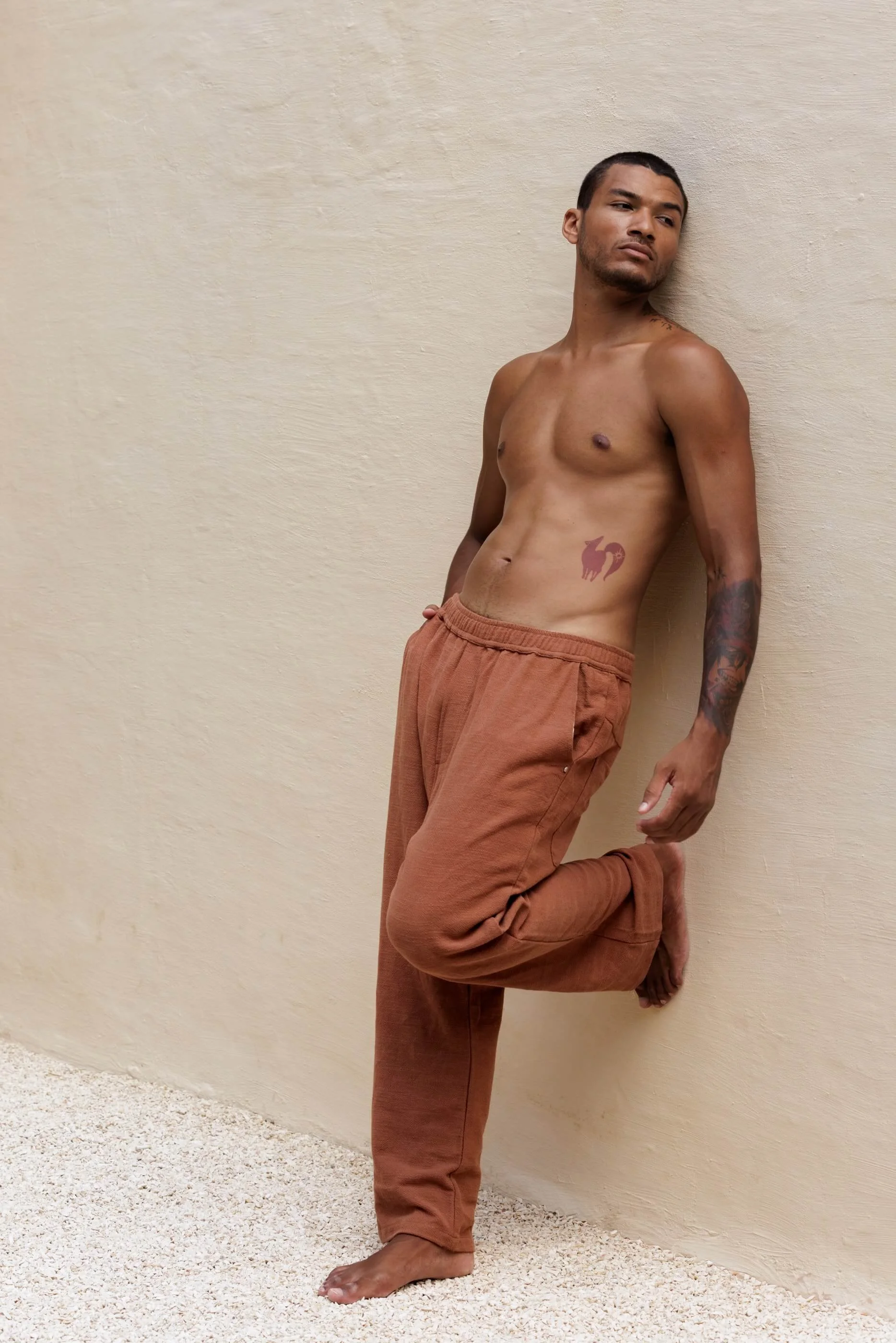 Full-length three-quarter front view of the model wearing the Benirras trousers in Terracotta, leaning against a wall to showcase the soft straight-leg linen silhouette.