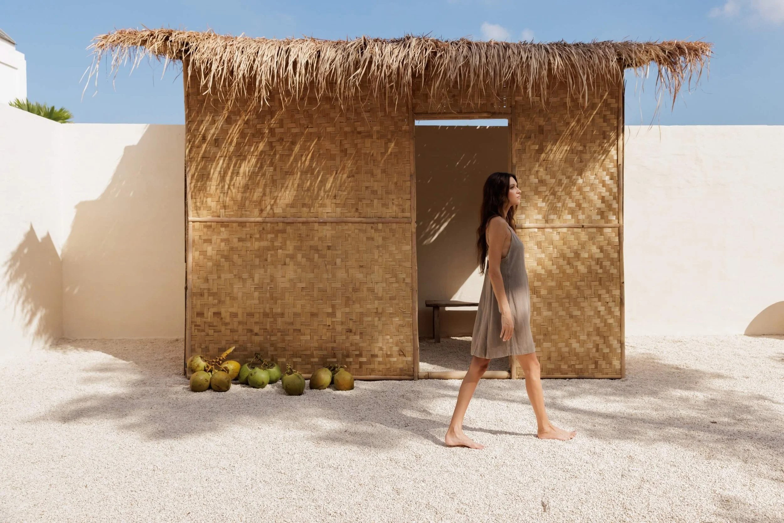 A woman in the NOUSA ISA dress in Linen Viscose bland, stone washed,  walking on sand in front of a woven bamboo shack, with green coconuts on the sand and a white wall on the left