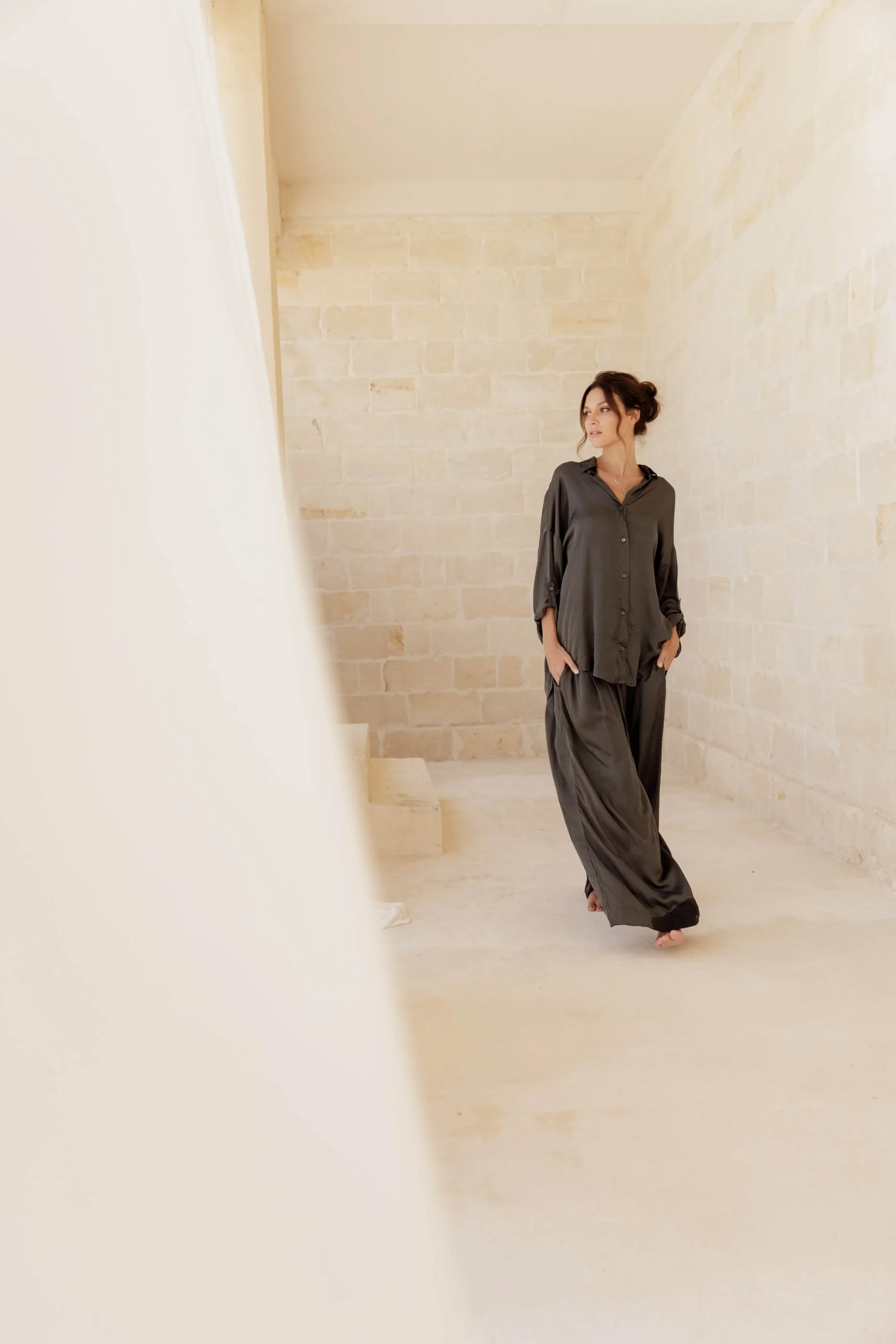 A distant editorial shot of the model walking, capturing the effortless elegance, liquid movement, and contemporary oversized silhouette of the Dust Grey satin shirt and pants ensemble.