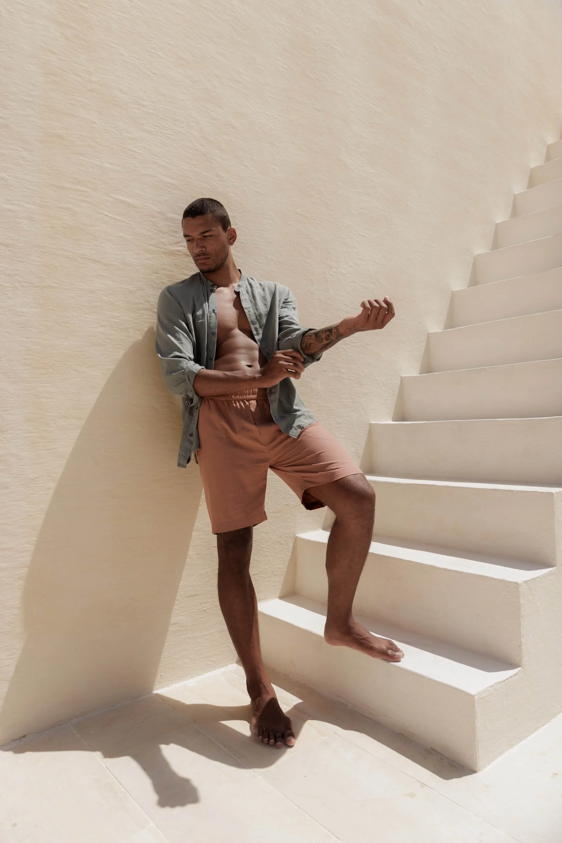 An editorial shot of the model at the base of white stairs against a white wall, wearing the Terra Stone shorts with the contrasting Sage Stone Jordi shirt left open.