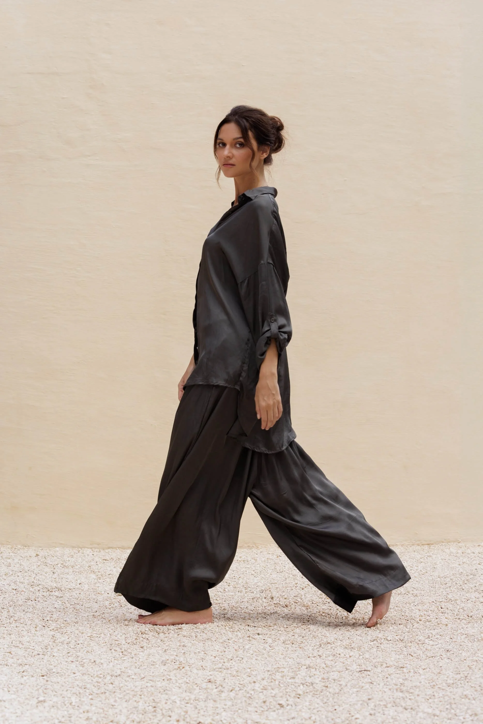 Side view of the model walking, showcasing the ultra-wide leg silhouette, fluid movement, and metallic sheen of the Dust Grey viscose satin Aguas pants.