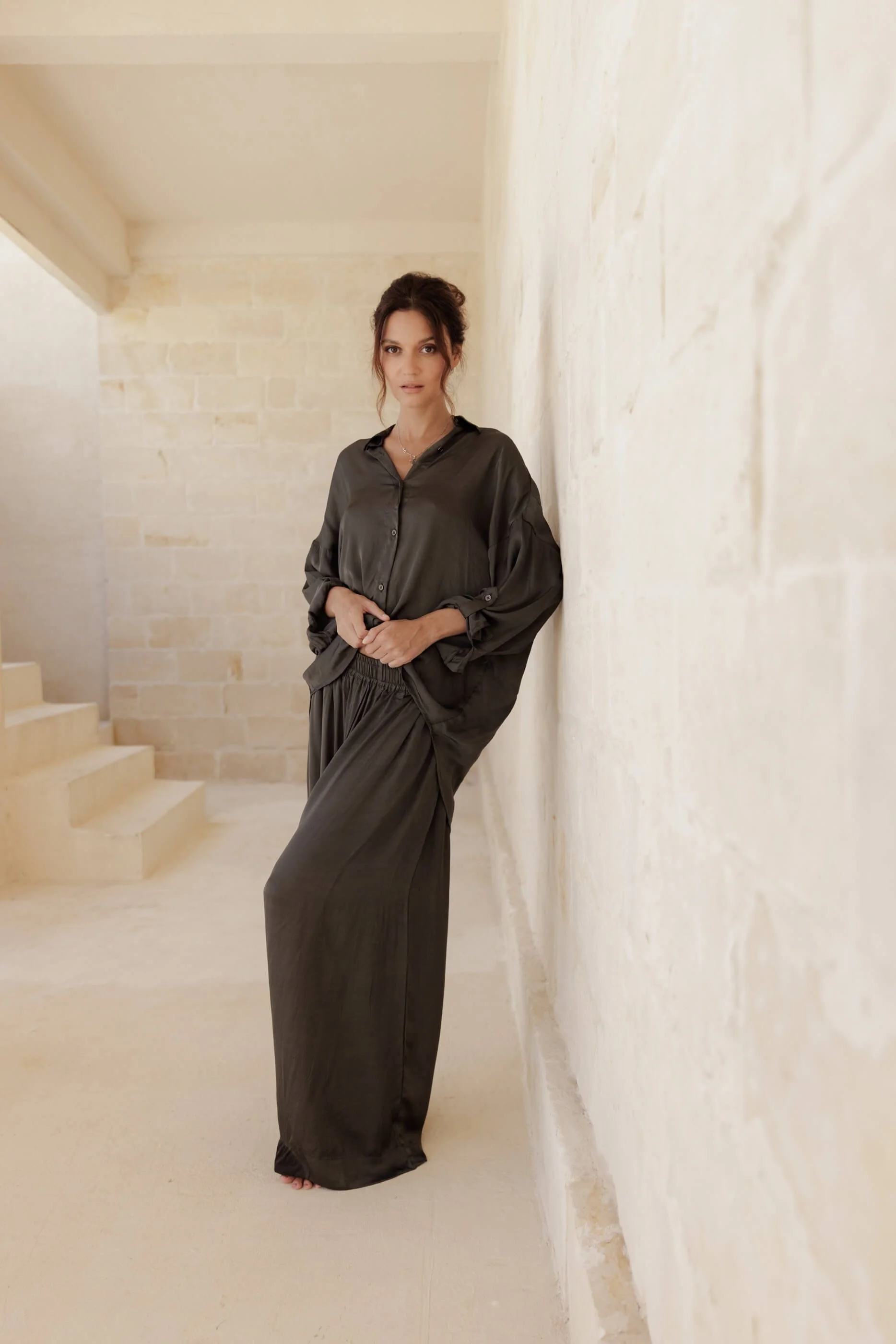 An editorial shot of the model leaning against a textured wall, showcasing the relaxed, fluid drape of the oversized Dust Grey Lolita shirt and matching wide Aguas pants.