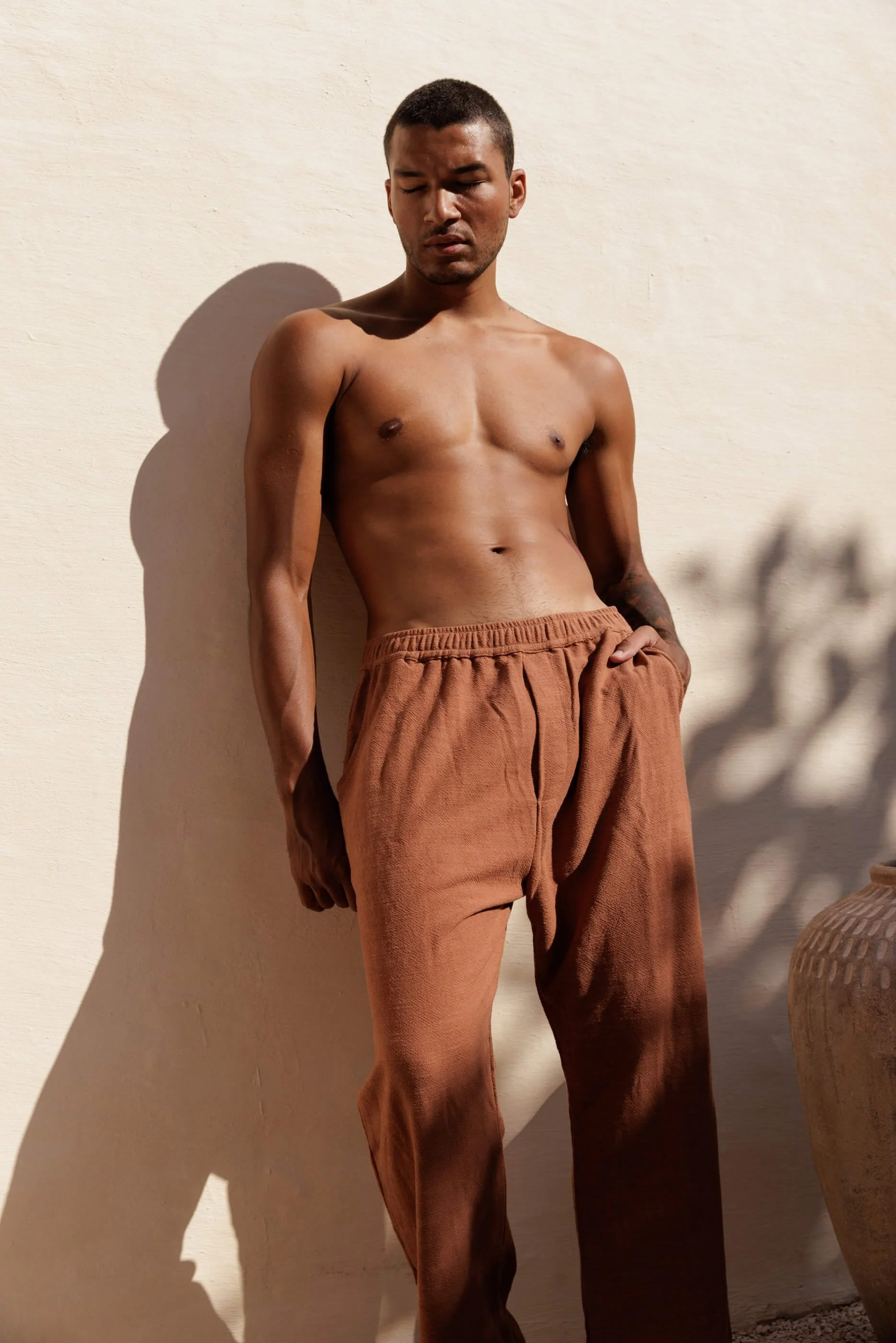 A centered, closer editorial shot of the model wearing the Terracotta linen trousers, leaning against the wall with soft shadows highlighting the rich, sun-baked brick orange texture.