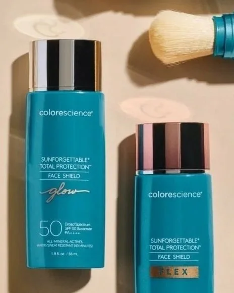 Two bottles of Colorescience Sunforgettable Total Protection Face Shield with SPF 50, one larger and one smaller, placed on a surface with a makeup brush nearby.