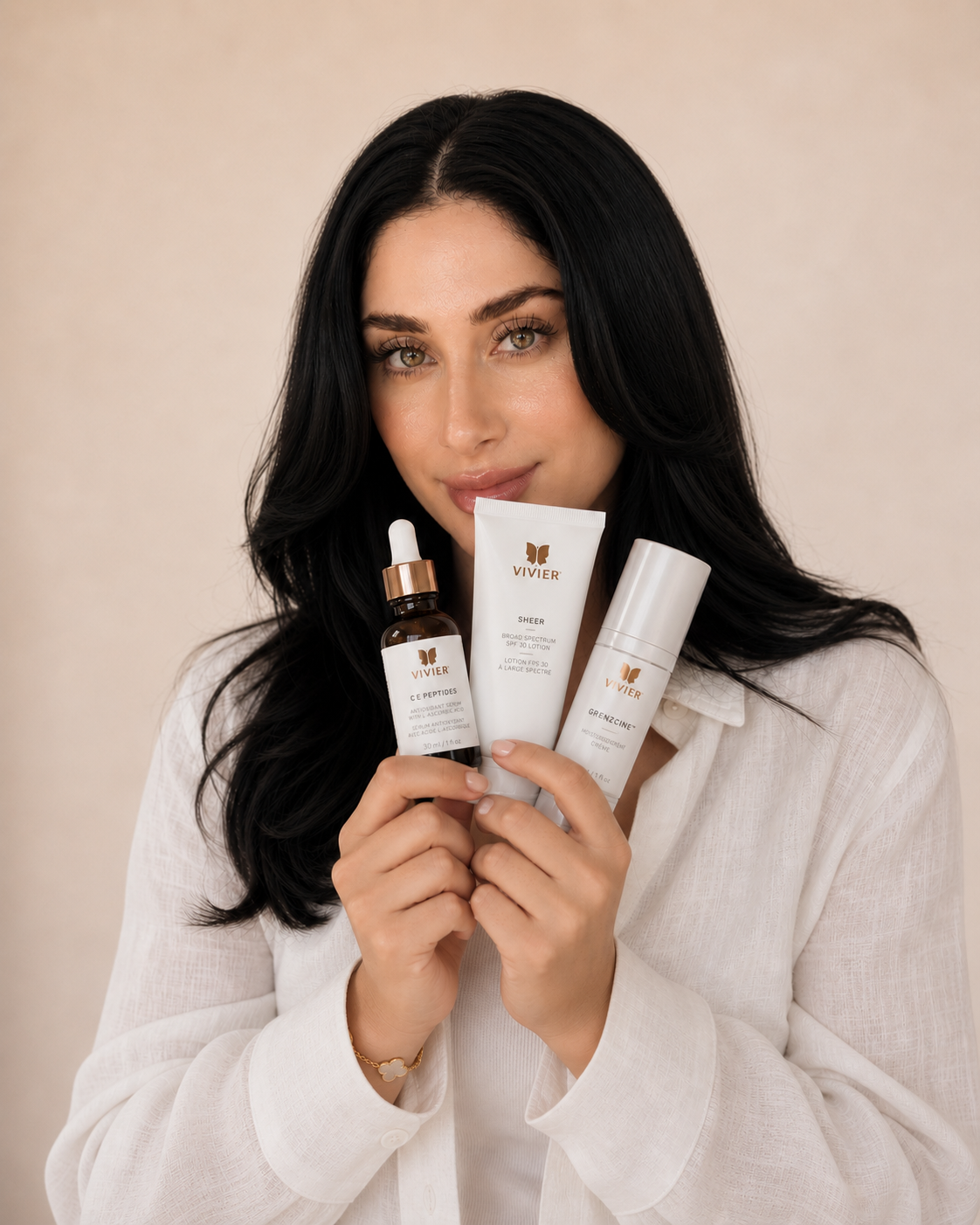 A woman with long black hair holding Vivier skincare products close to her face, looking at the camera with a slight smile.