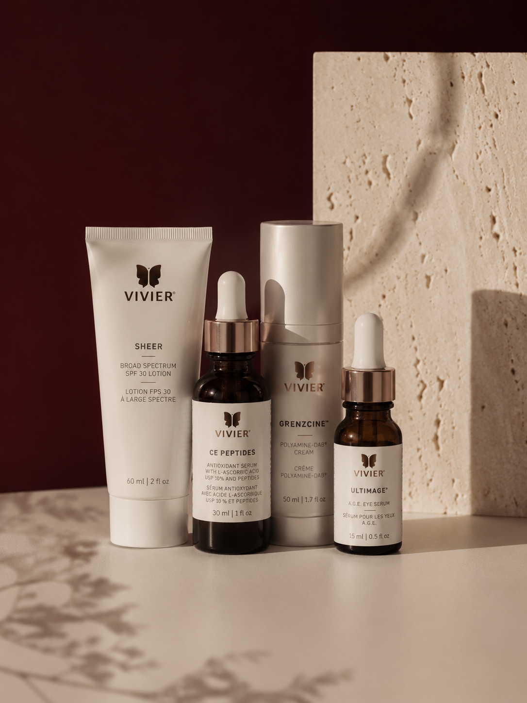 Collection of skincare products, including VIVIER Sheer Broad Spectrum SPF 30 lotion, CE Peptides antioxidant serum, GrenzCine polyamine-DAB cream, and UltimAge A.G.E. eye serum, arranged on a surface with a textured stone background and a dark red wall.