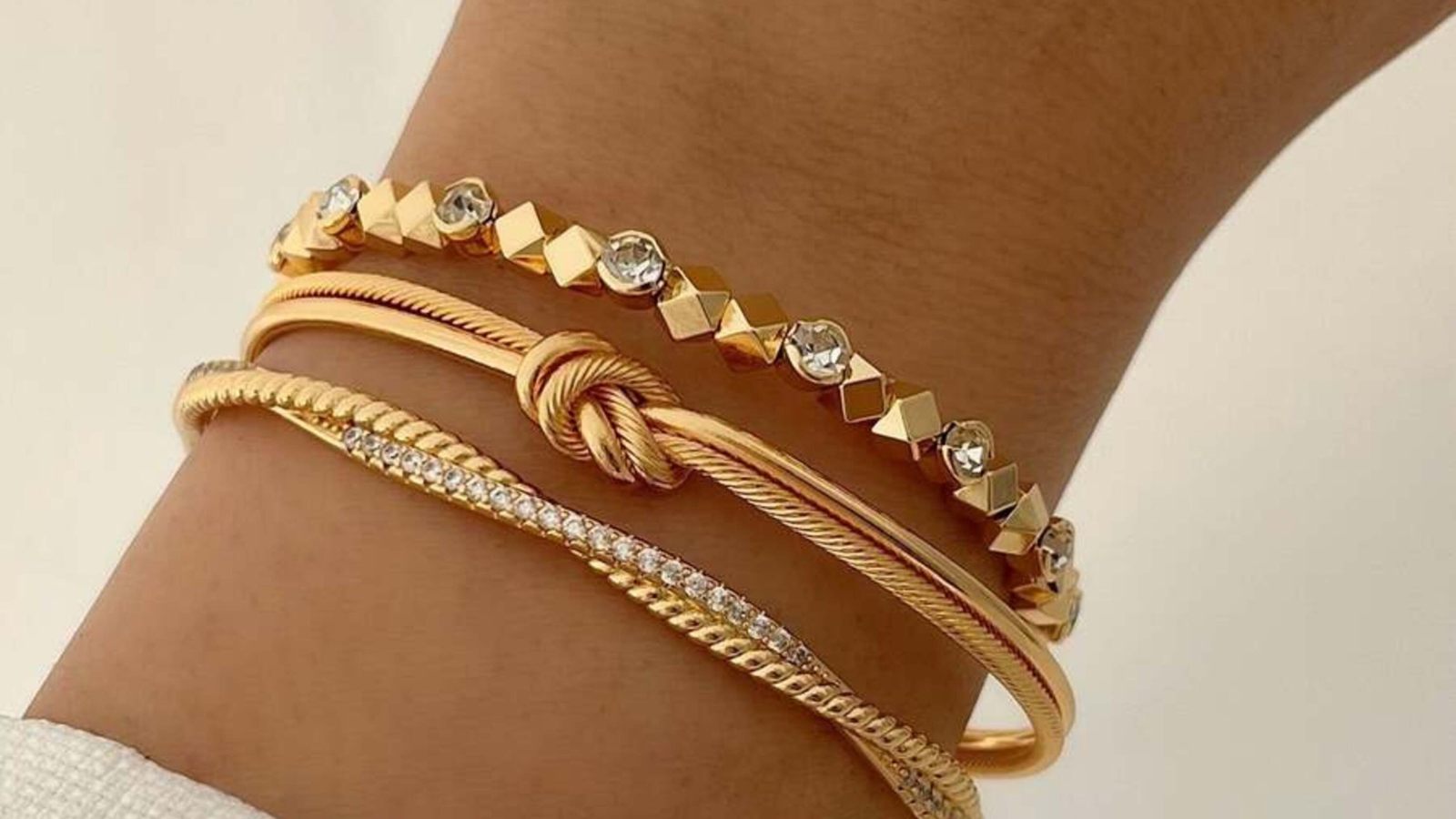 Three gold bracelets on a person's wrist, including a geometric studded bracelet, a twisted rope bracelet with a knot, and a thin diamond-studded chain bracelet.