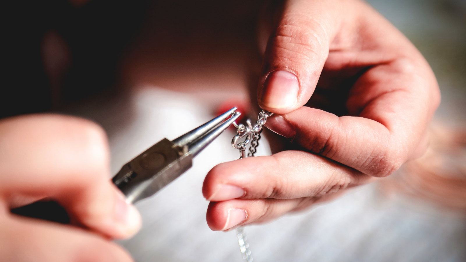 Close-up of hands holding jewelry pliers and working on a silver chain necklace.