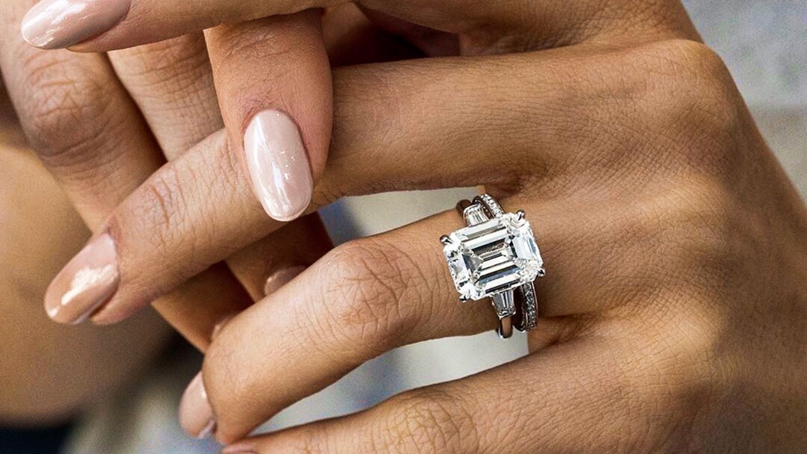 Close-up of two hands with rings, one hand has a large emerald-cut diamond engagement ring and an eternity band, both with metal bands; the other hand has a polished nude-colored manicured nails, touching the ring.