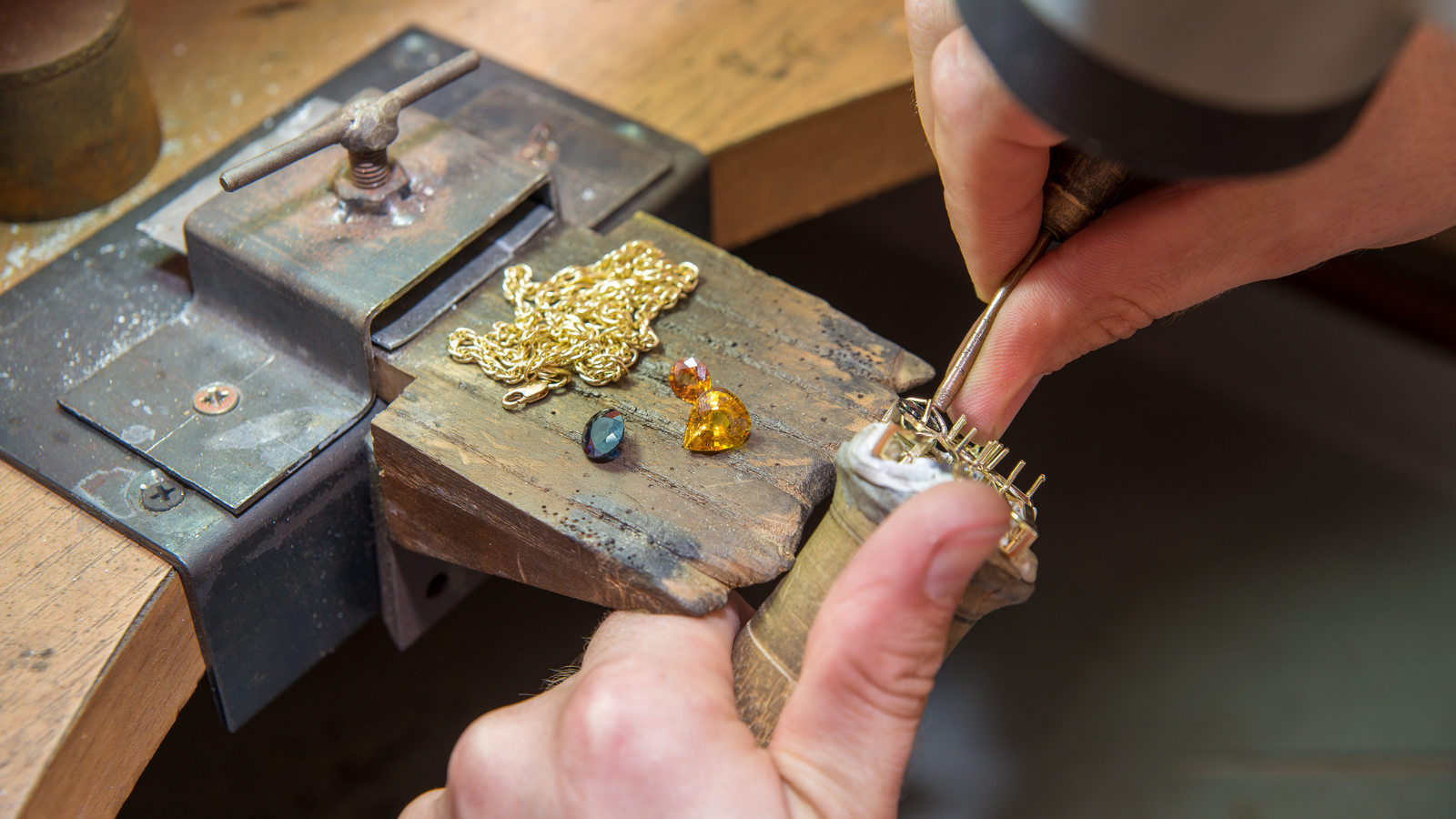 Jeweler working on a necklace using a small precision tool at a workbench, with jewelry pieces and gemstones nearby.