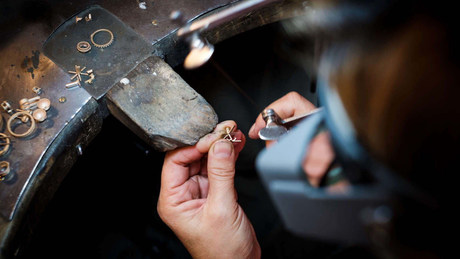 A person working on jewelry, using pliers to shape or assemble a small metal piece at a workbench with various small metal components around.