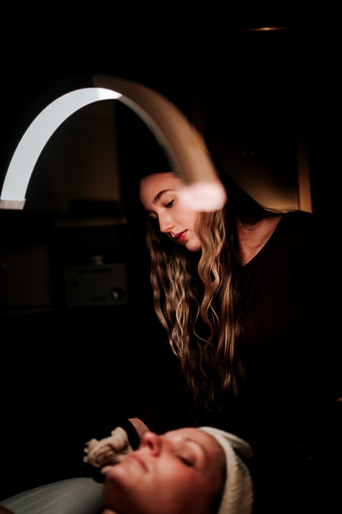A woman with long wavy hair looks down at a dog, which is lying on a person's chest with closed eyes, in a dimly lit room.