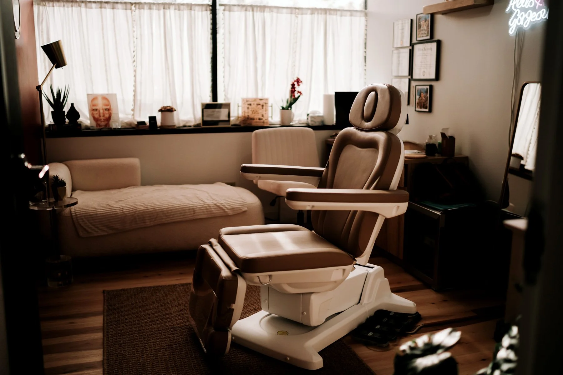 A medical examination room featuring a reclining exam chair, a beige couch with a blanket, a window with curtains, and framed certificates on the wall.