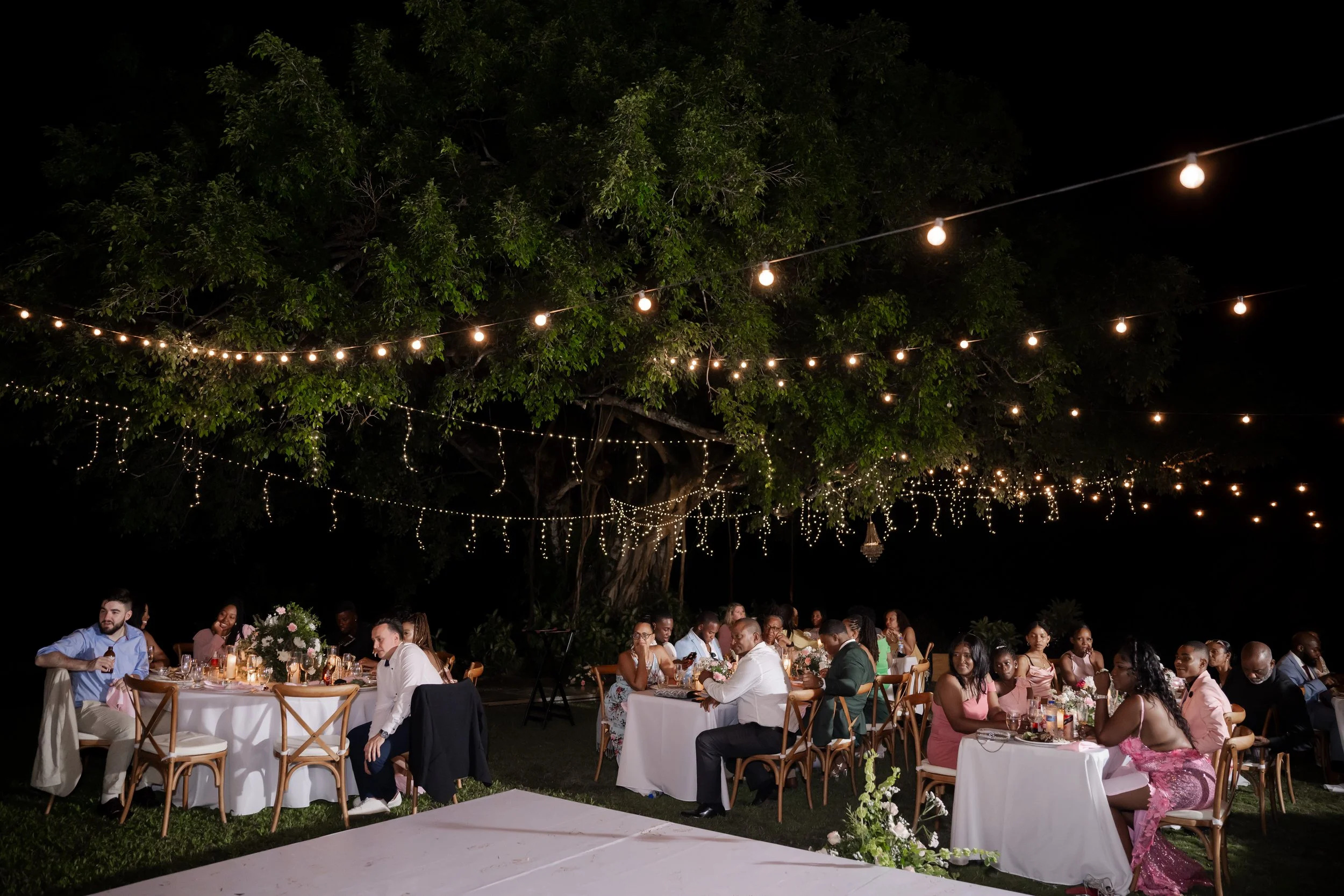 Outdoor nighttime celebration with guests sitting at tables, string lights hanging from trees, and a dark sky above.
