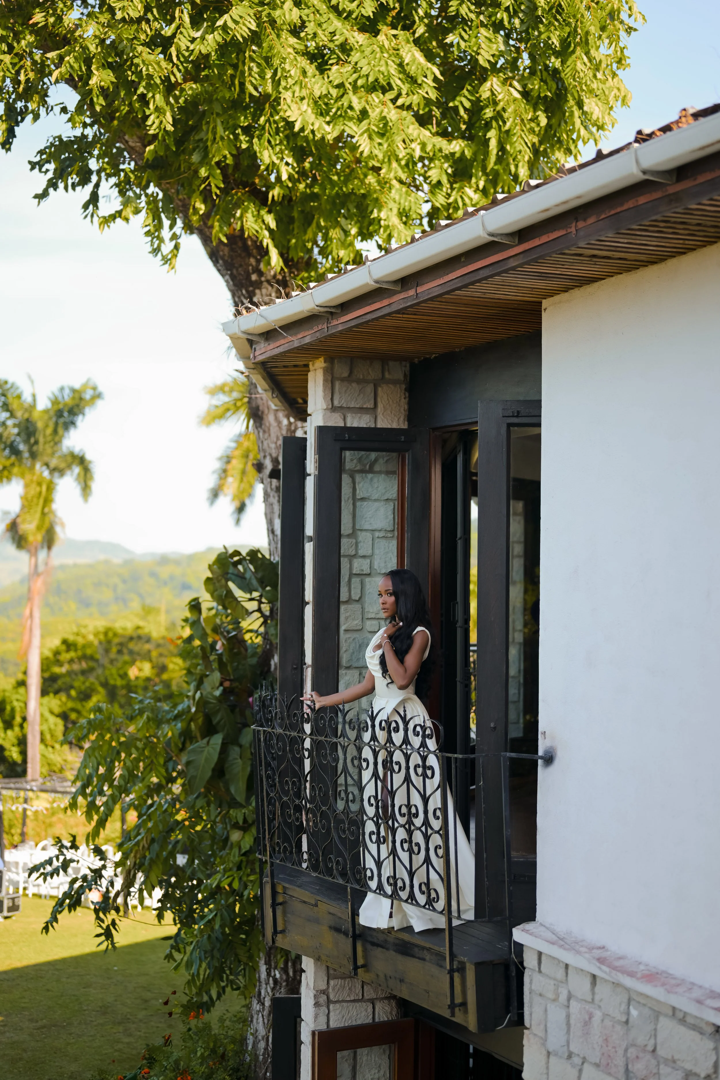 A woman with long black hair standing on a balcony of a house, looking outside. The house has a stone and white wall with black window frames and a decorative black iron railing. There are green trees and a clear sky in the background.