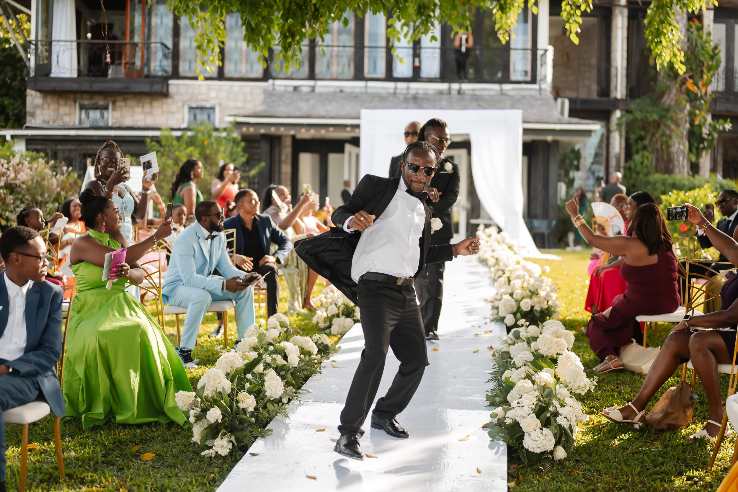 A man in a tuxedo and sunglasses is dancing on a white aisle at an outdoor wedding ceremony. Guests seated on either side of the aisle are watching and taking photos. The setting is decorated with white flowers and greenery with a multi-story house i