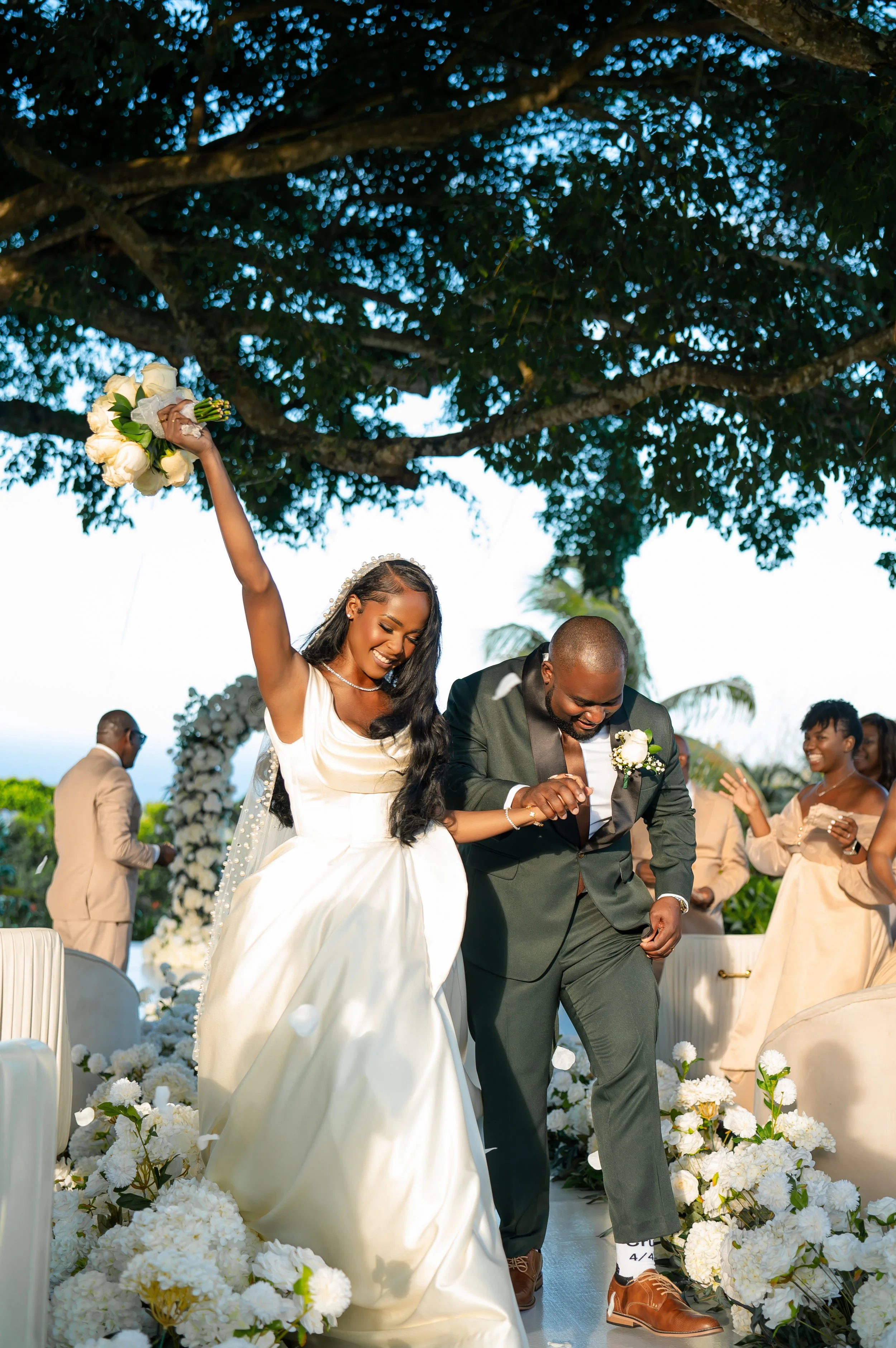 A bride and groom celebrating during their wedding ceremony outdoors, surrounded by white flowers, trees, and wedding guests.