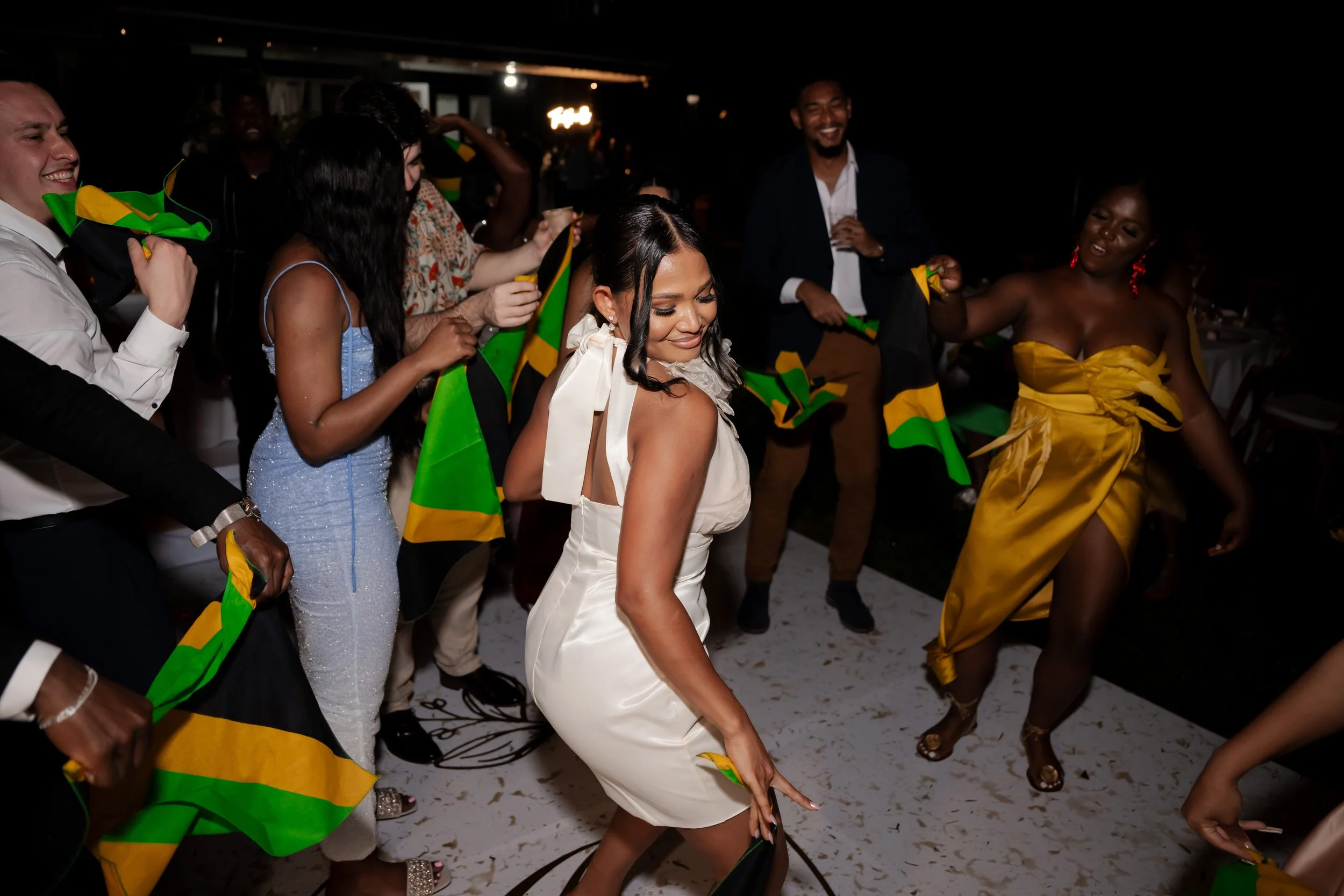 People dancing at a celebration, some holding Jamaican flags, with a woman in a white dress in the center.