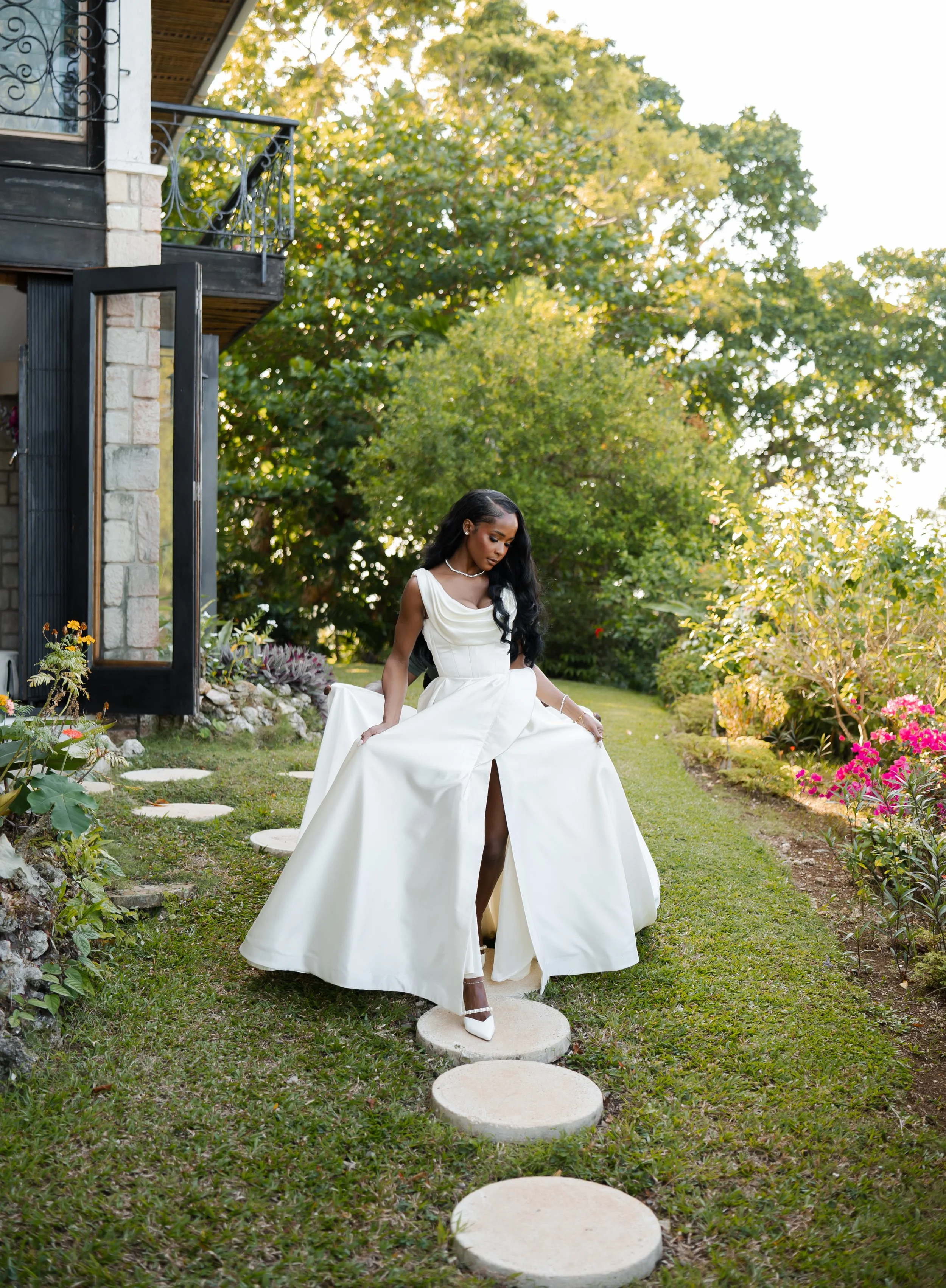 A woman in a white wedding dress standing on a stone pathway in a garden with green trees and colorful flowers, during daytime.