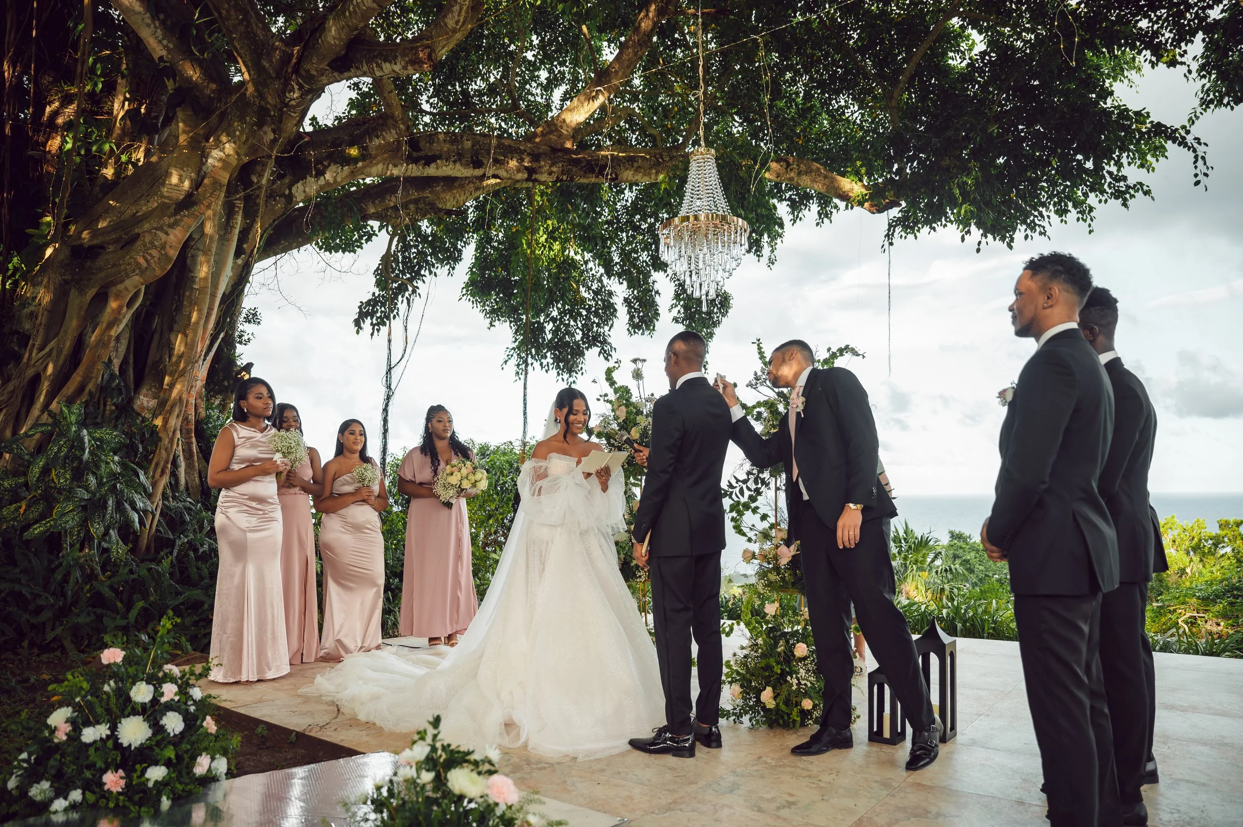 A wedding ceremony taking place outdoors beneath a large tree. The bride and groom are standing at an altar, with bridesmaids and groomsmen nearby, dressed in formal attire. The bride is wearing a white wedding gown, and the groom is in a dark suit. 