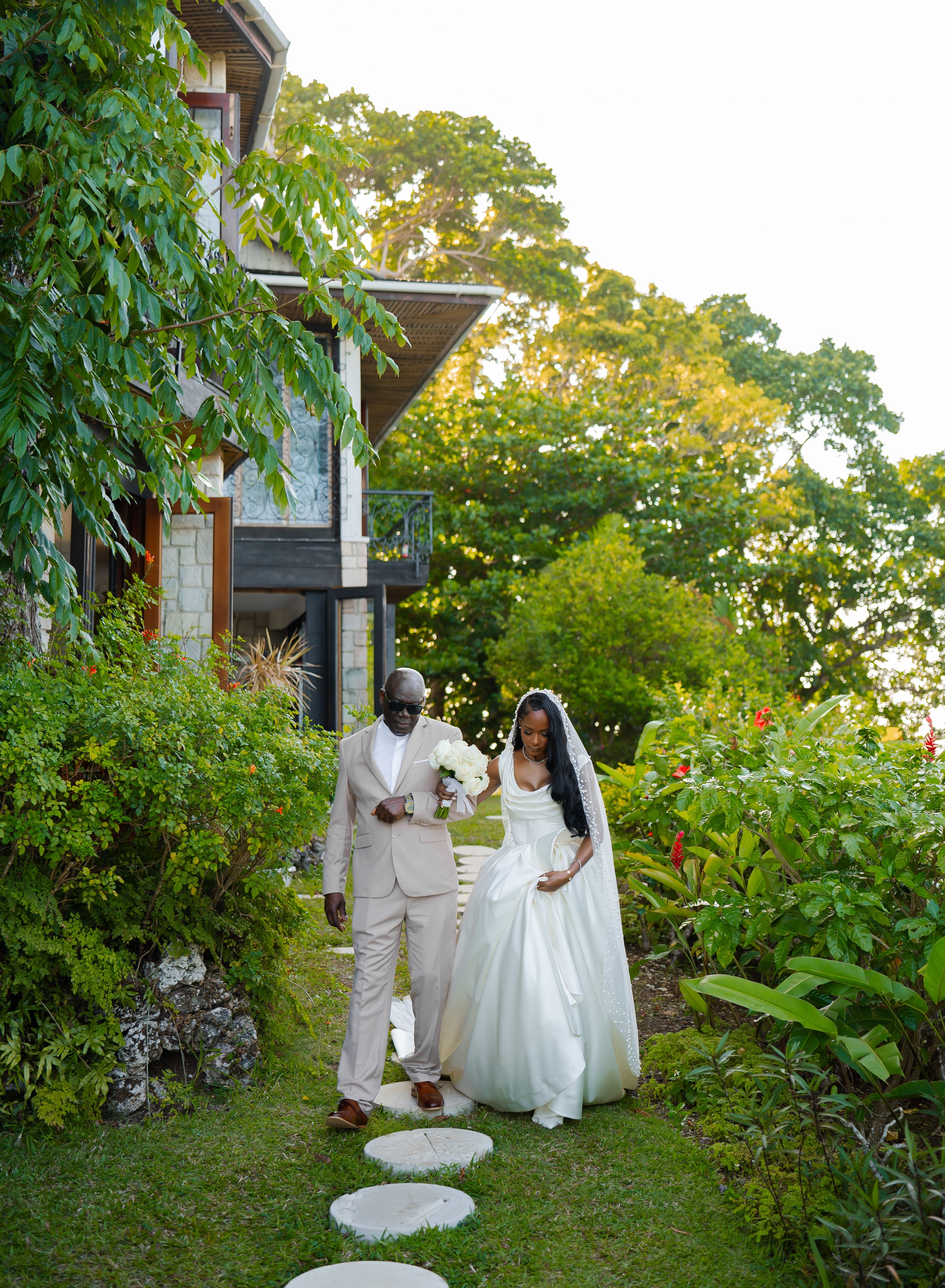 A man and a woman, dressed in wedding attire, walking down a garden path lined with lush green bushes and colorful flowers, with a house in the background.