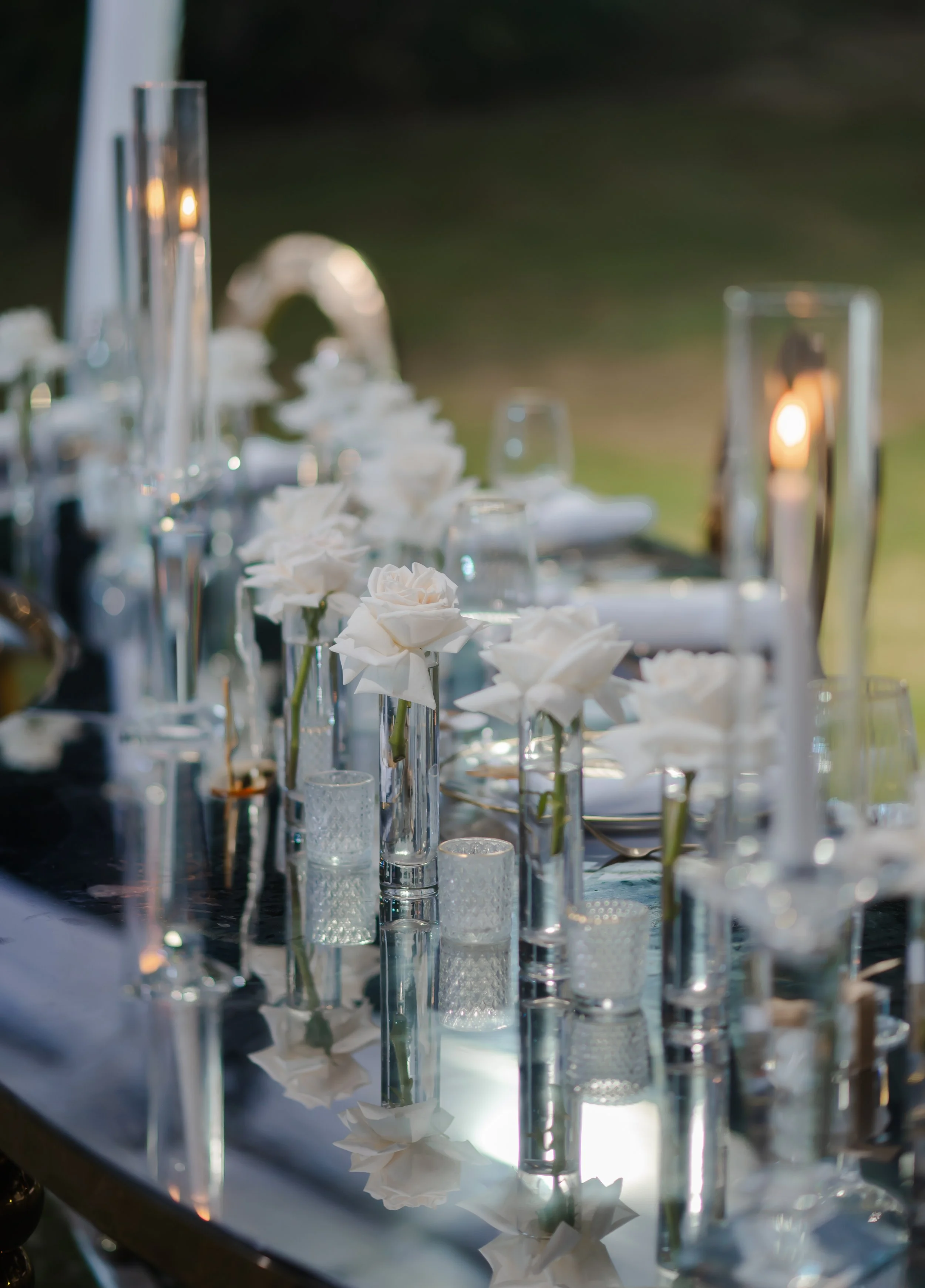 Elegant outdoor dining table decorated with white roses in glass vases, candles, and glassware, with candles lit, during what appears to be sunset or evening.