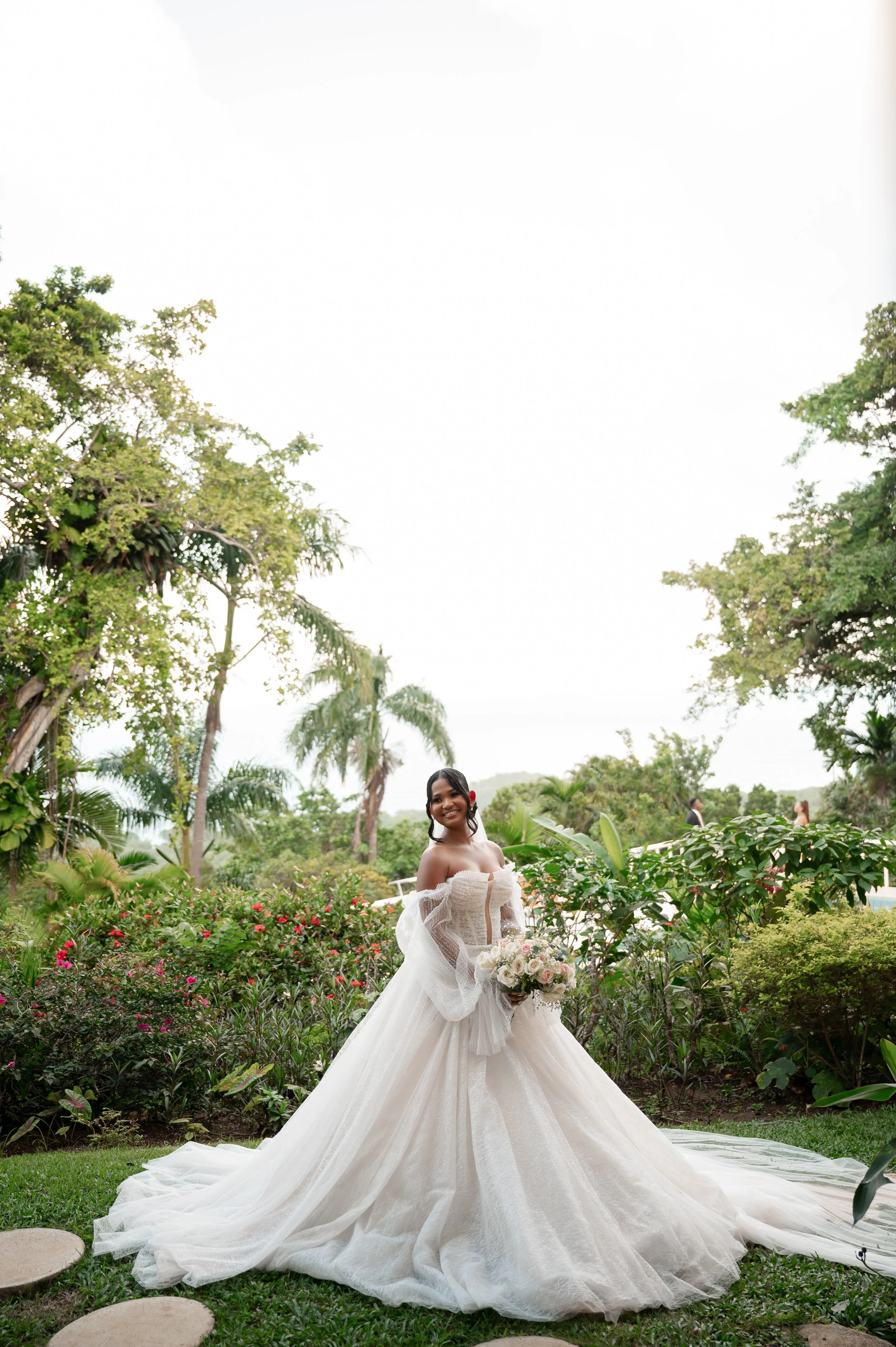 A bride in a strapless white wedding gown holding a bouquet of pink and white roses, standing in a lush garden with green trees and flowering bushes.