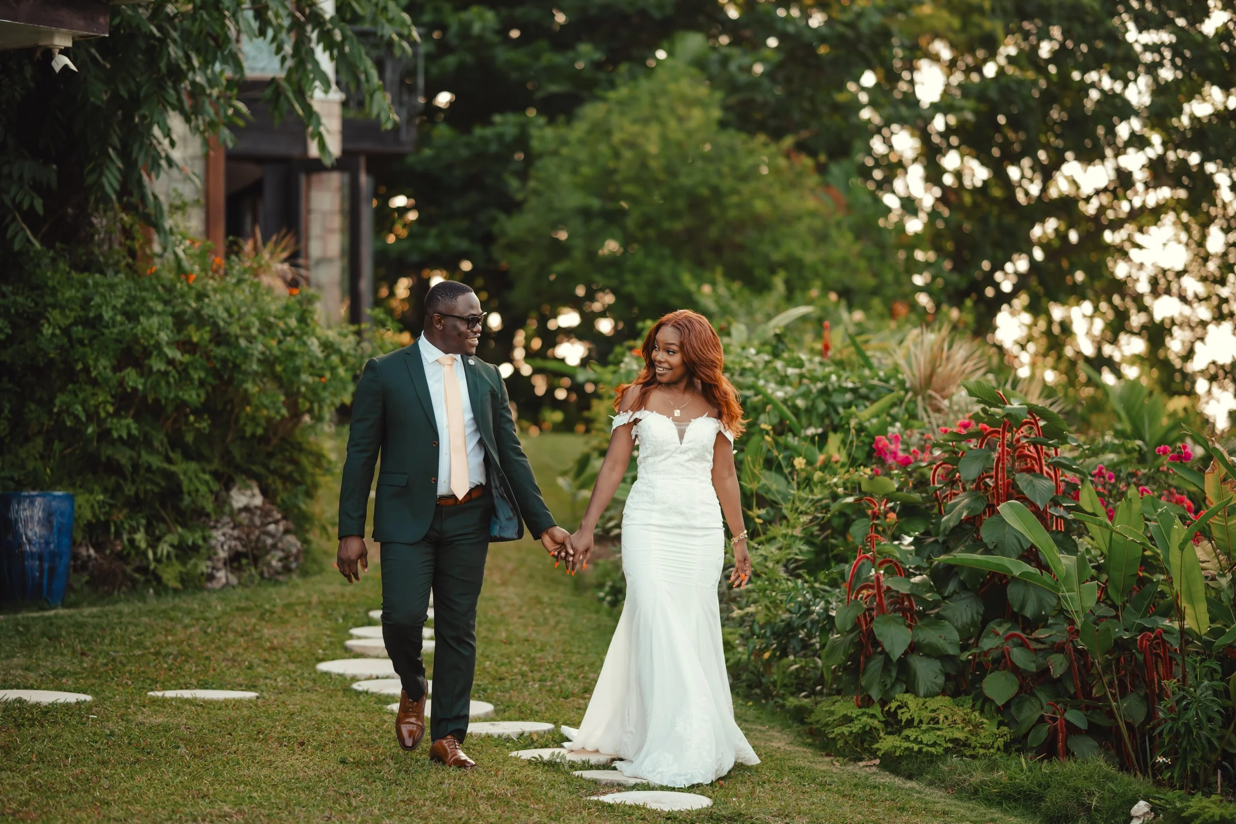 A newlywed couple walking hand in hand through a garden with lush green trees and colorful flowers during the daytime.