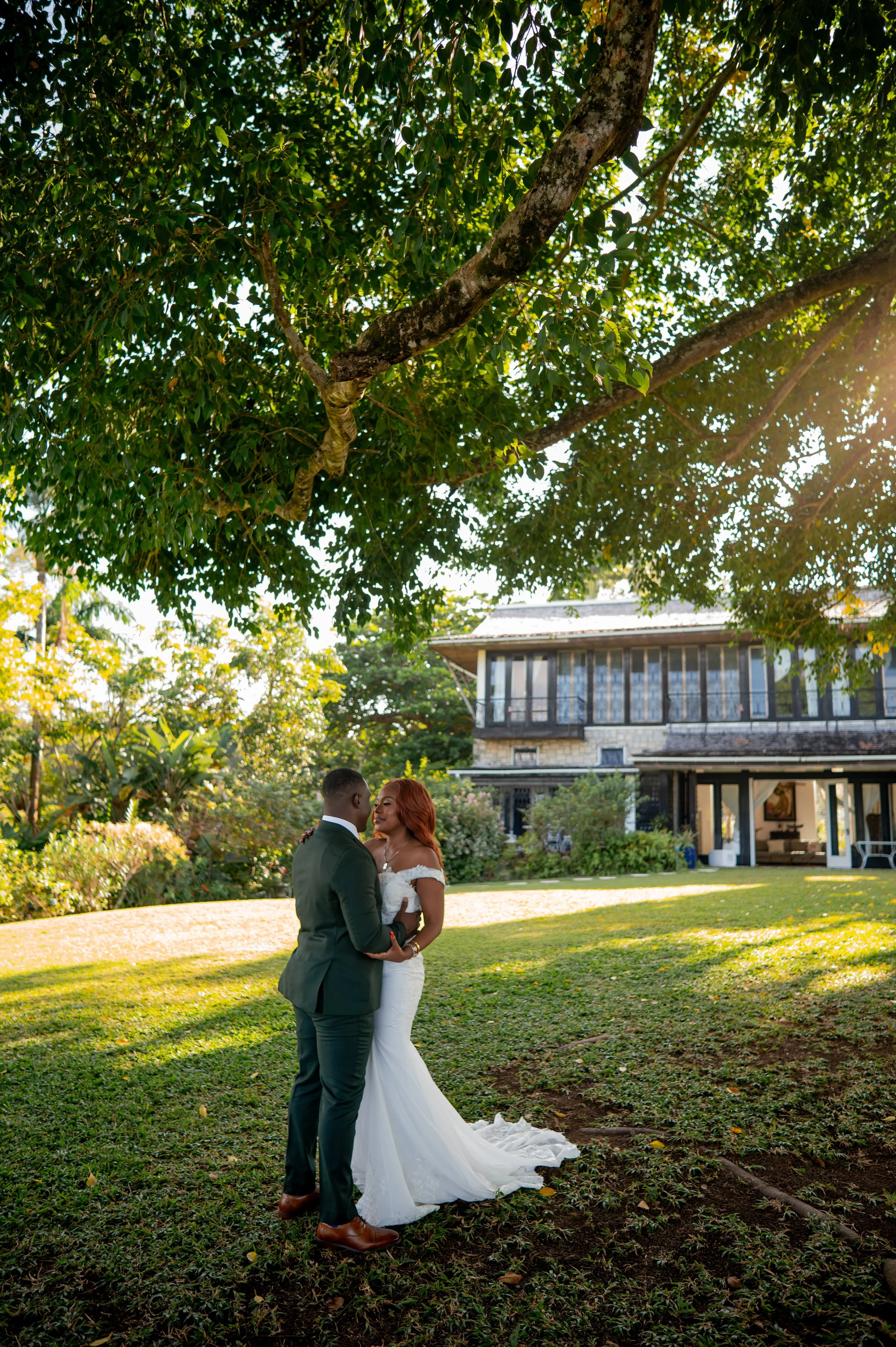 A bride and groom standing in a lush green garden, gazing lovingly at each other. The bride wears a white wedding gown and the groom a dark suit. They are surrounded by trees and sunlight filters through the leaves, with a house visible in the backgr