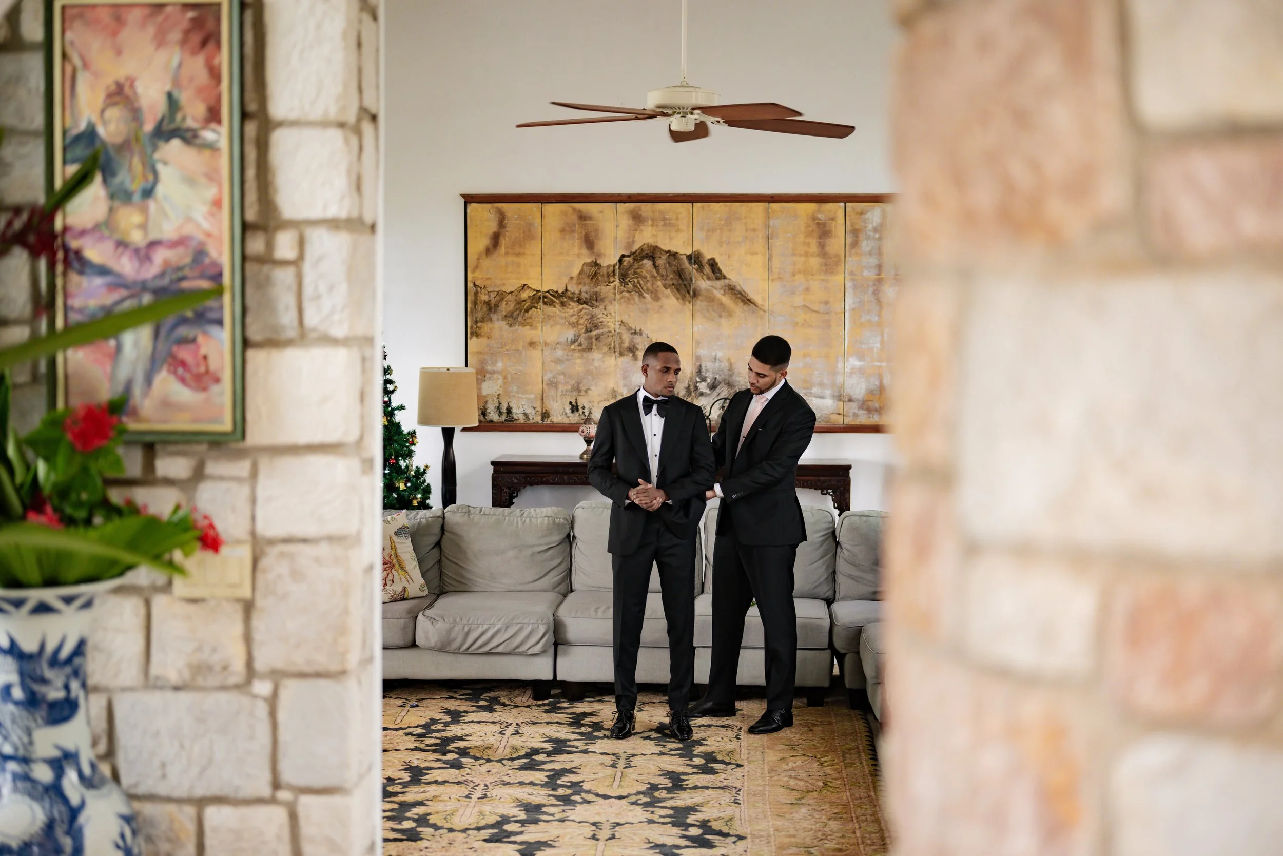Two men dressed in black tuxedos standing together in a living room, possibly preparing for a formal event. The background features a large floral and mountain landscape artwork, a beige sofa, a decorated Christmas tree, a modern lamp, and a patterne