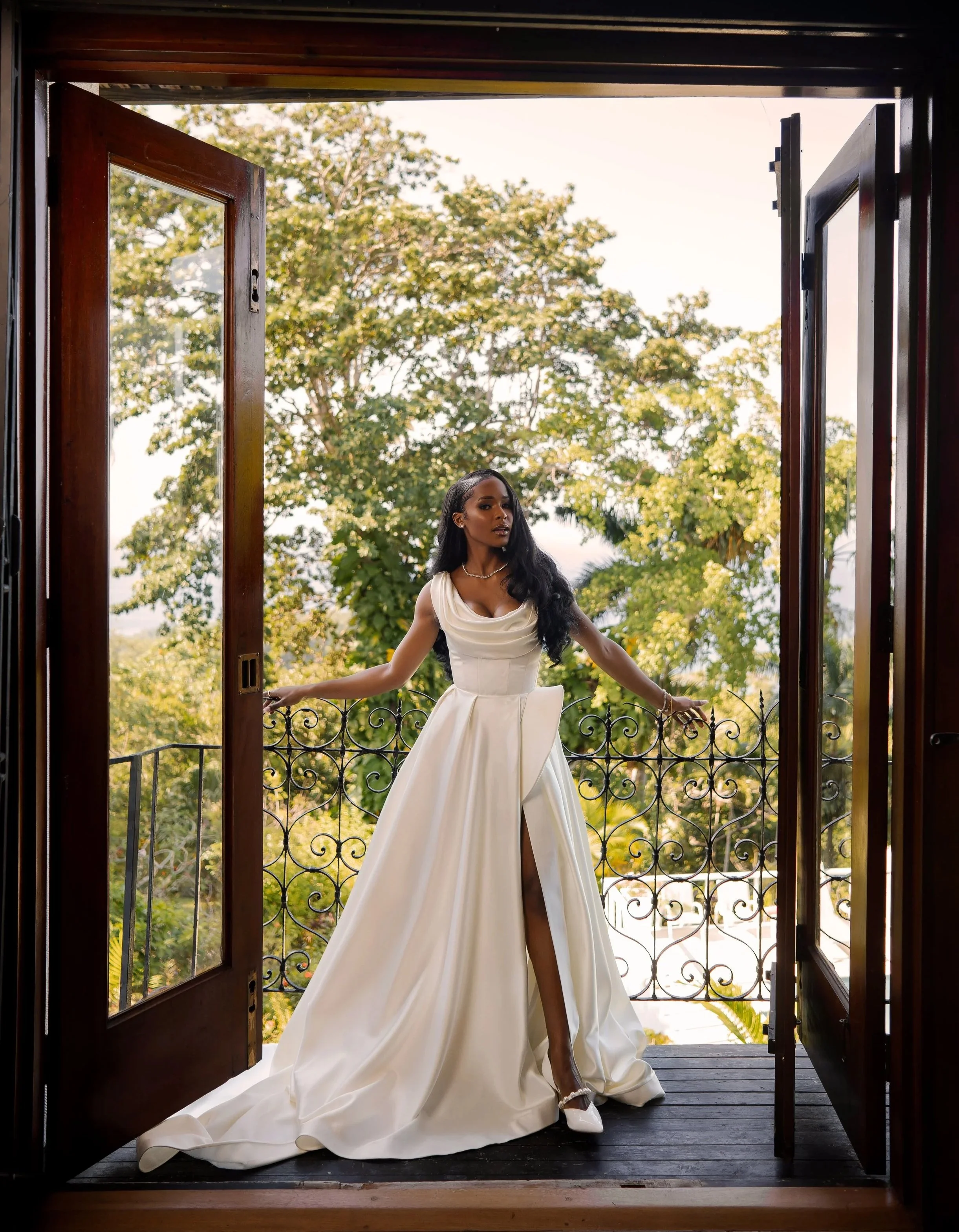 A woman in a white wedding dress standing at a balcony with open wooden doors and a black wrought iron railing, surrounded by green trees.