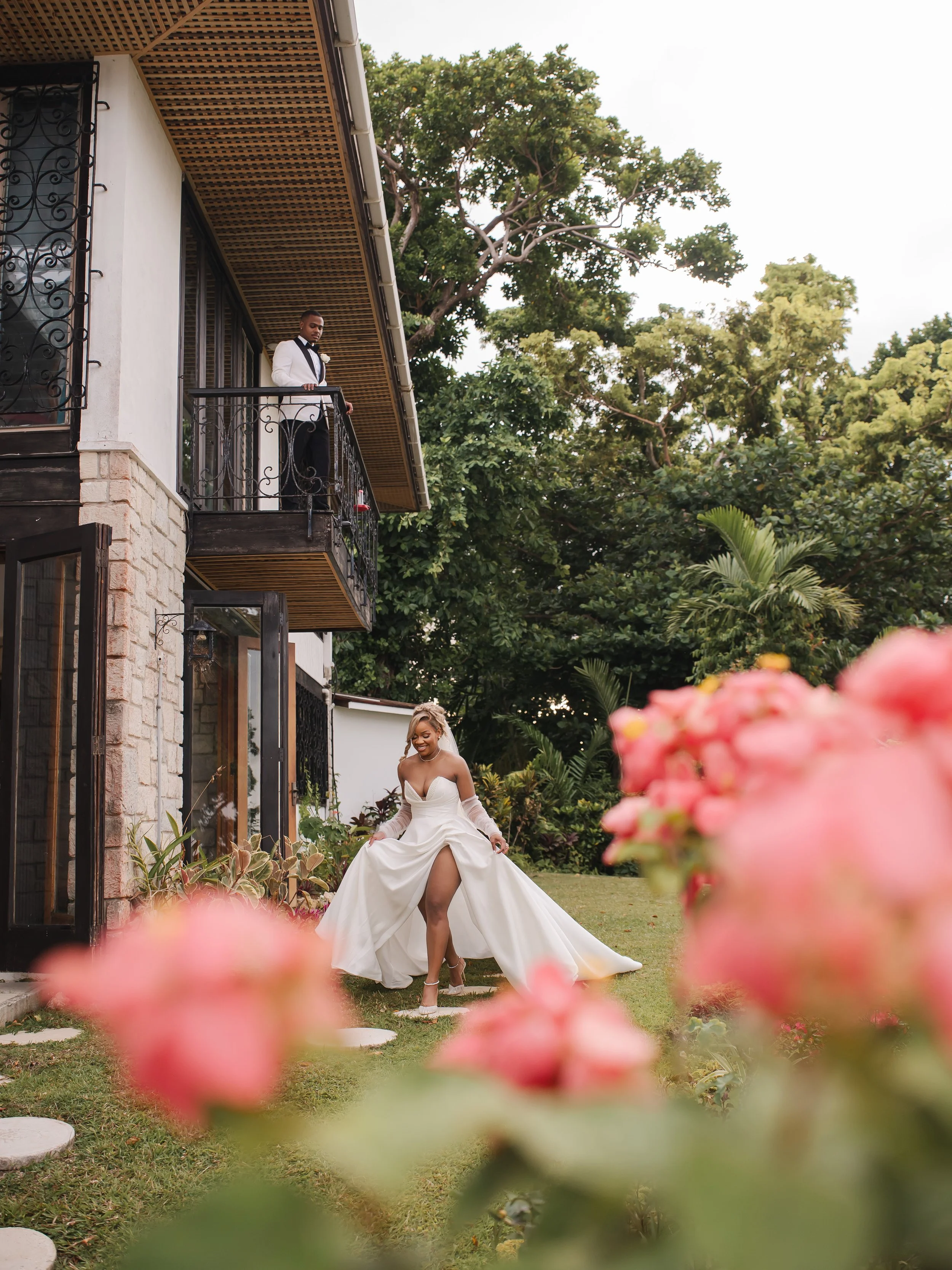 A bride in a white wedding dress with a slit, standing in a garden with pink flowers in the foreground. A man in a white tuxedo with black accents is on a balcony above, looking down at her.