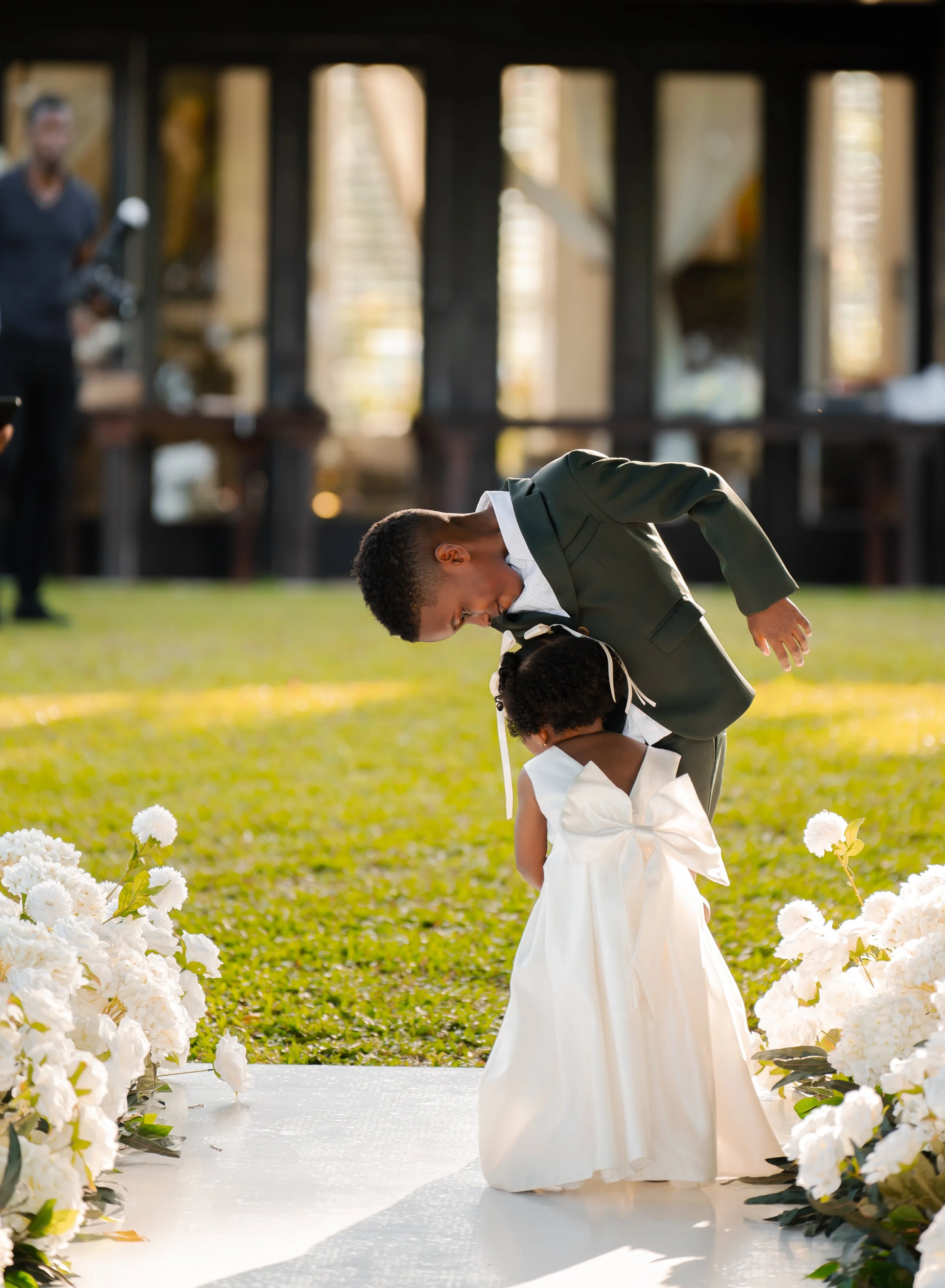 A young boy in a formal suit kneeling and bowing during a wedding ceremony, with a woman in a wedding dress kneeling before him, surrounded by white flowers on either side and greenery in the background.