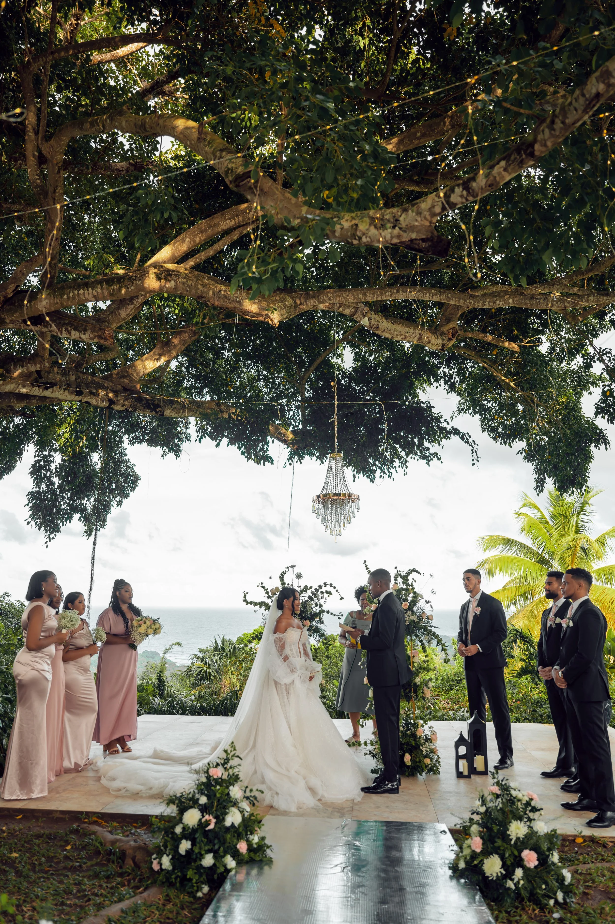 Outdoor wedding ceremony with bride and groom exchanging vows under a large tree decorated with string lights. Bridesmaids and groomsmen stand on either side, floral arrangements and lanterns are visible, ocean view in the background.