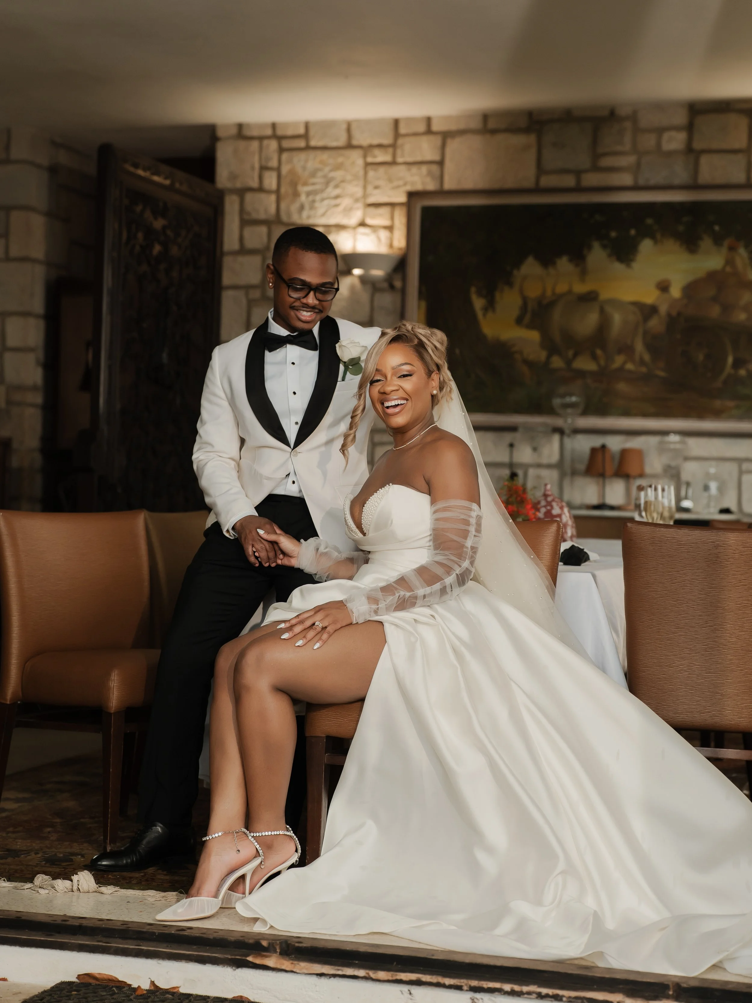 A bride and groom on their wedding day, sitting at a reception, smiling and enjoying each other's company, with the bride wearing white heels and the groom in a tuxedo.