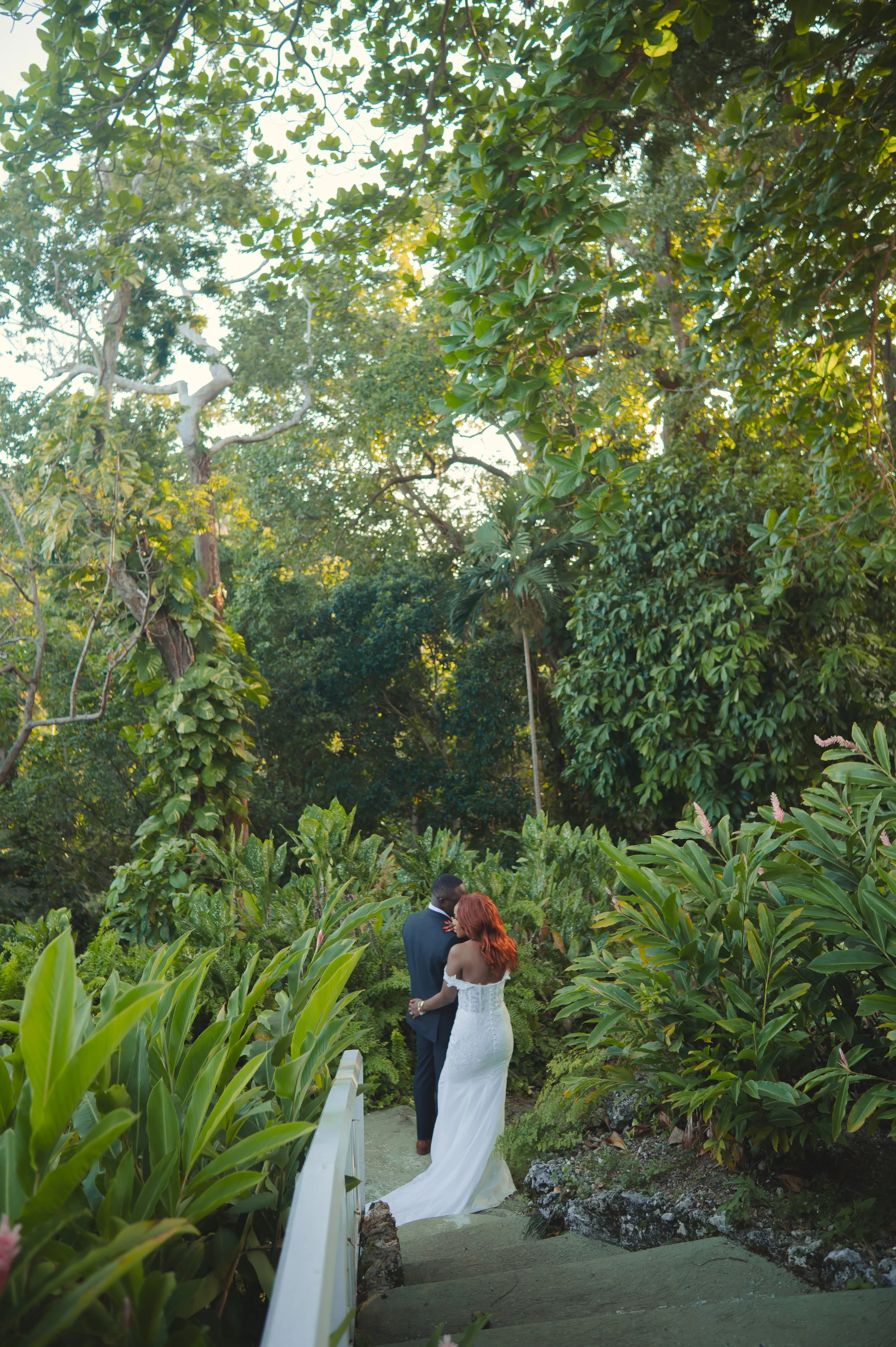 A bride and groom standing on a lush green garden pathway surrounded by dense trees and plants, sharing an intimate moment during their wedding.