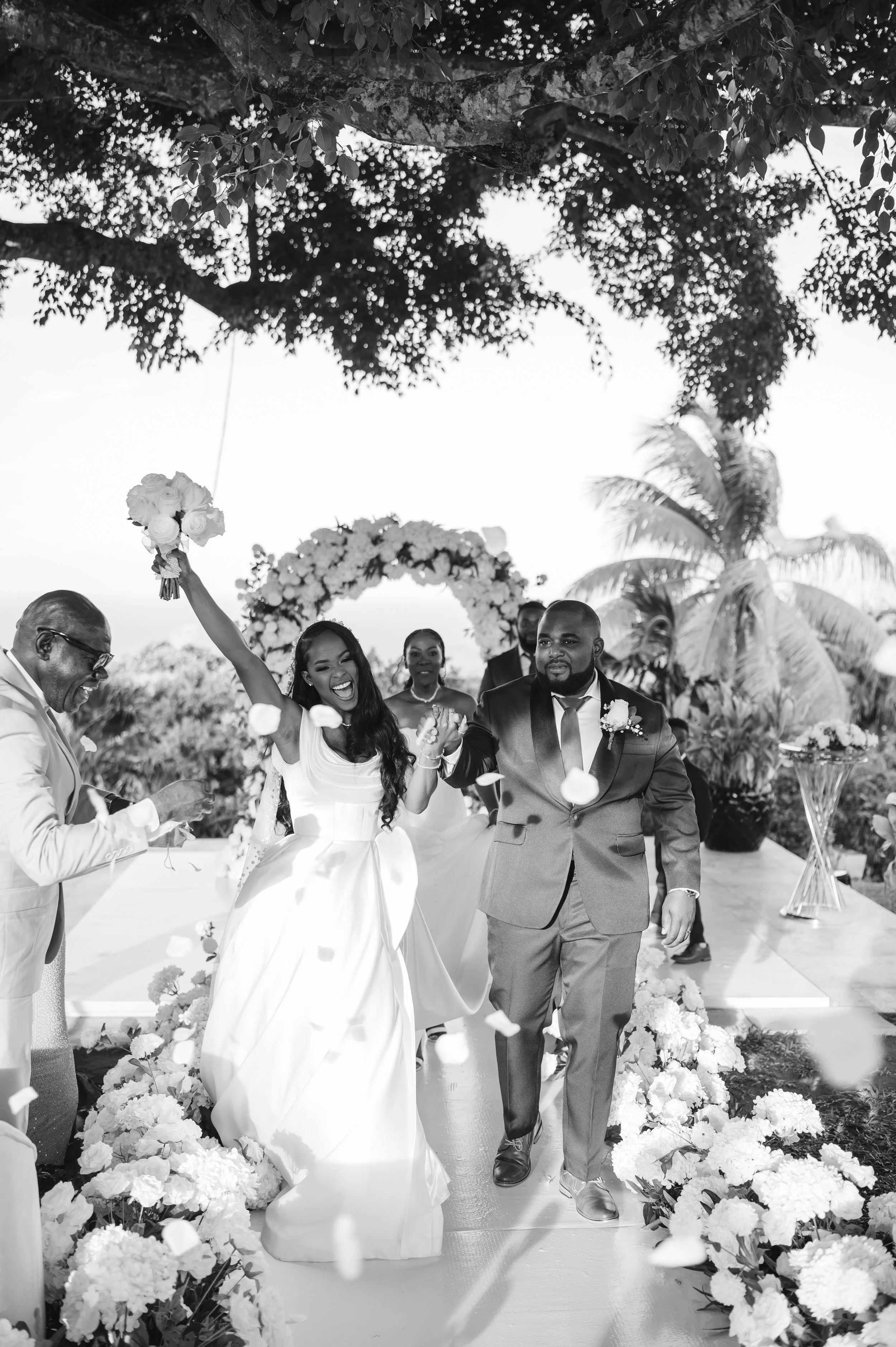 Black and white photo of a wedding ceremony outdoors, with a bride and groom holding hands and smiling, surrounded by guests and floral decorations.