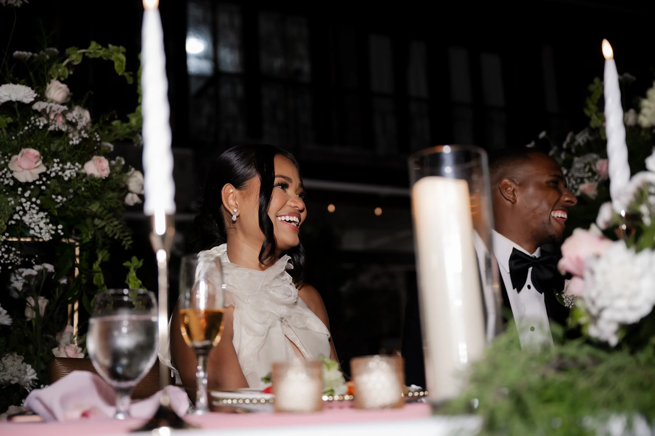 A smiling woman with black hair and earrings sitting at a wedding reception table decorated with flowers, candles, and glassware, with a man in formal tuxedo beside her. Flowers and candles adorn the background.