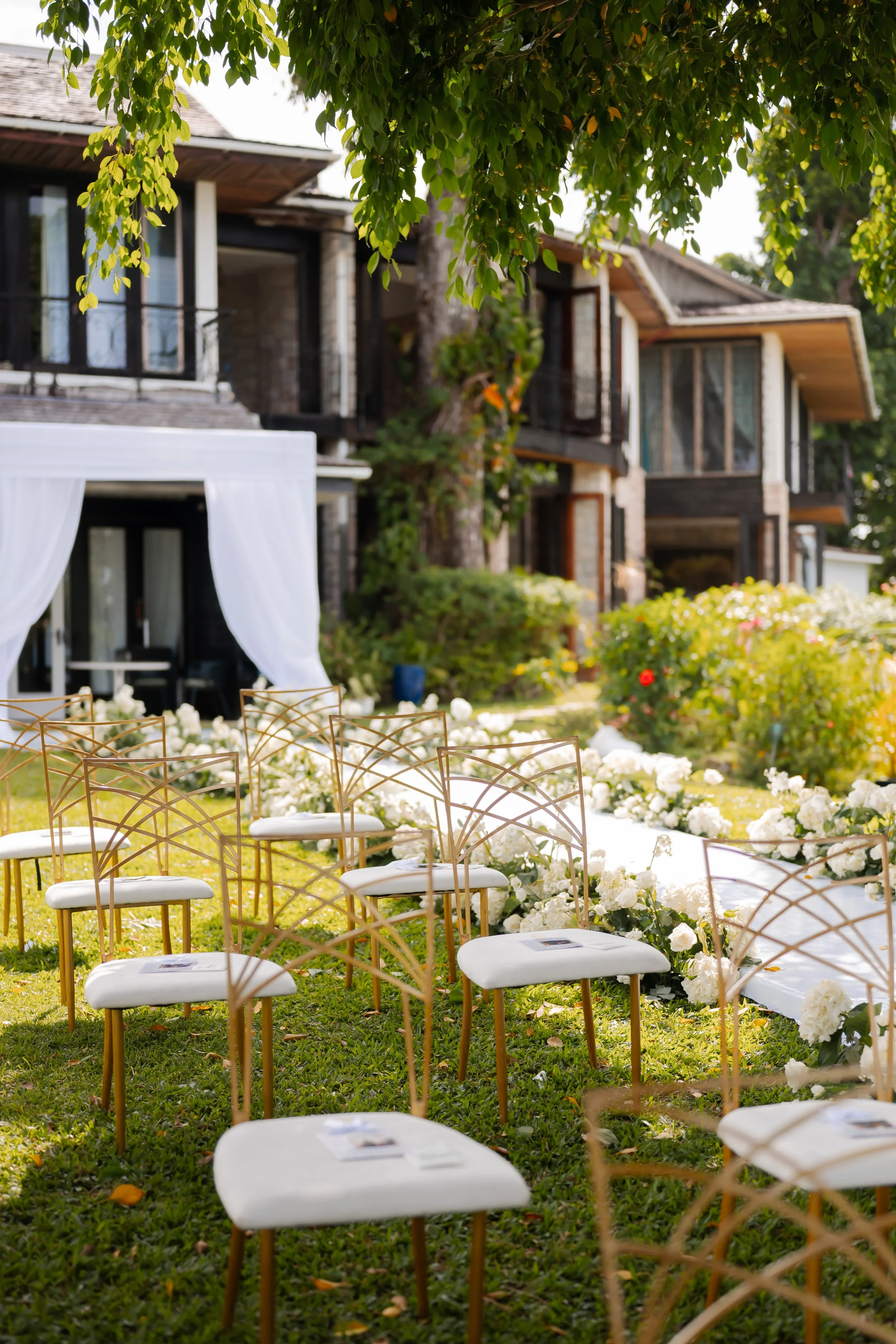 Outdoor wedding setup with white chairs, white flower decorations, a pathway lined with white flowers, and a building with large windows and balconies in the background surrounded by green trees.