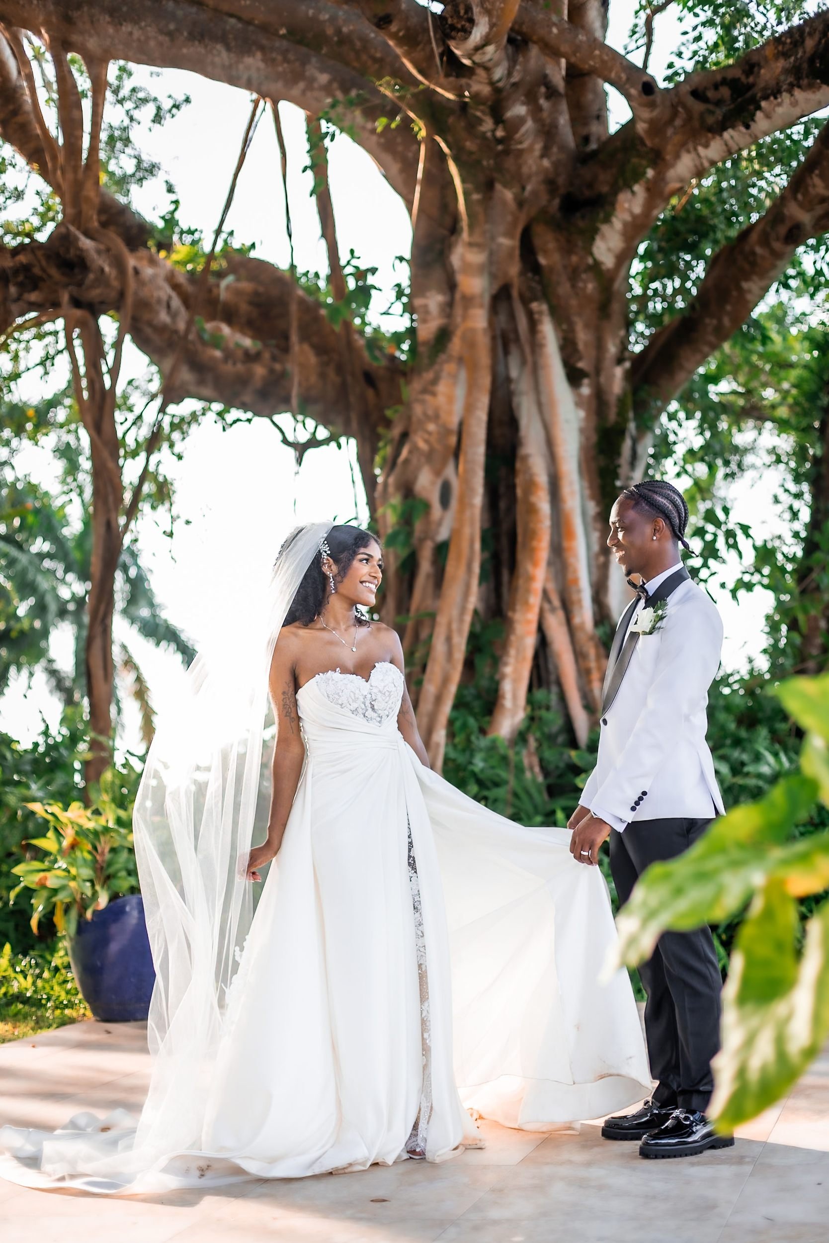 A bride and groom standing outdoors underneath a large tree, facing each other during their wedding ceremony. The bride is wearing a strapless white wedding gown with a lace bodice and a long veil, while the groom is dressed in a white tuxedo jacket, black pants, and a black bow tie.