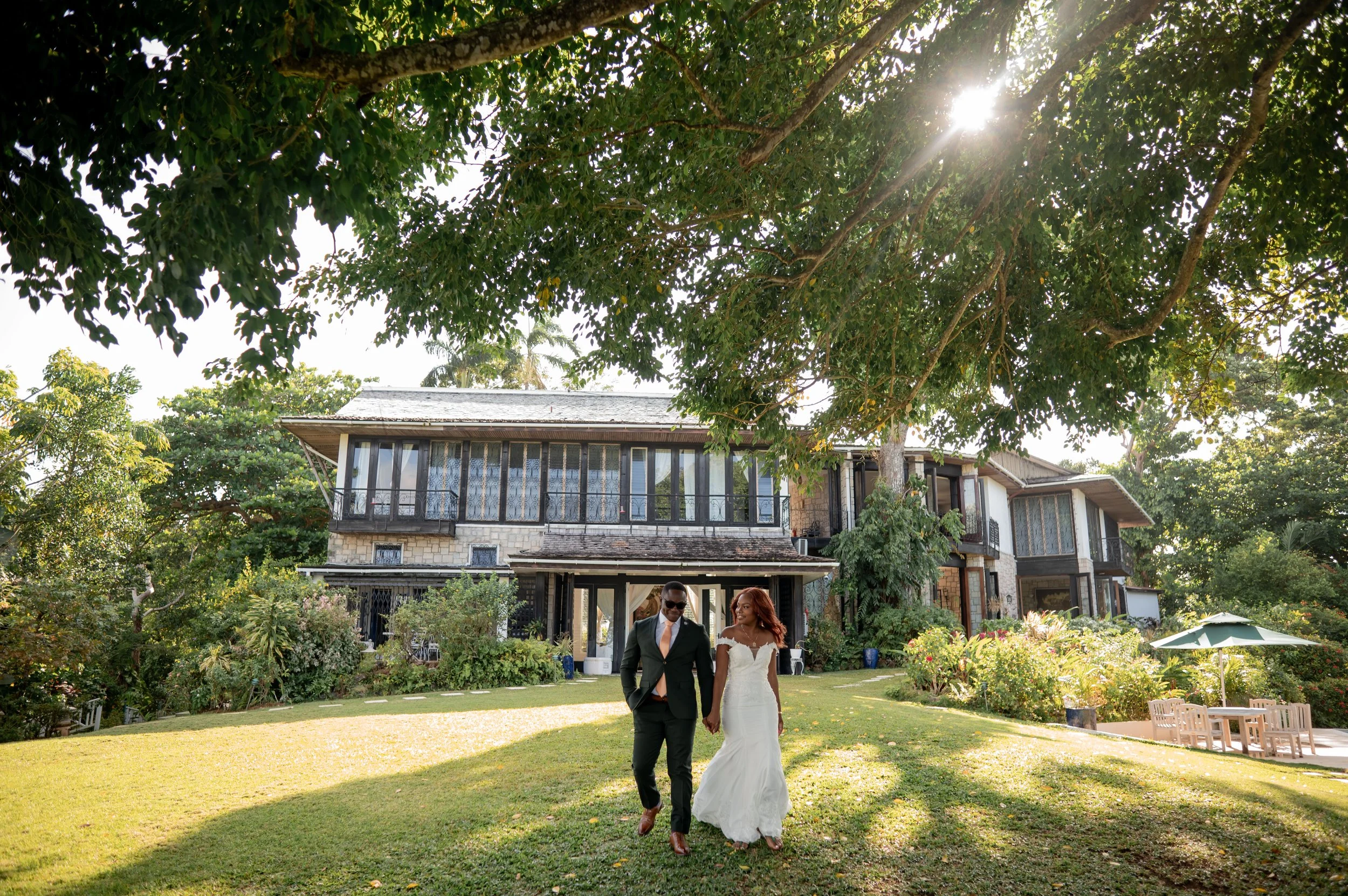 A newlywed couple walking arm in arm across a lush green lawn in front of a large house with greenery and trees, with sunlight filtering through the leaves overhead.