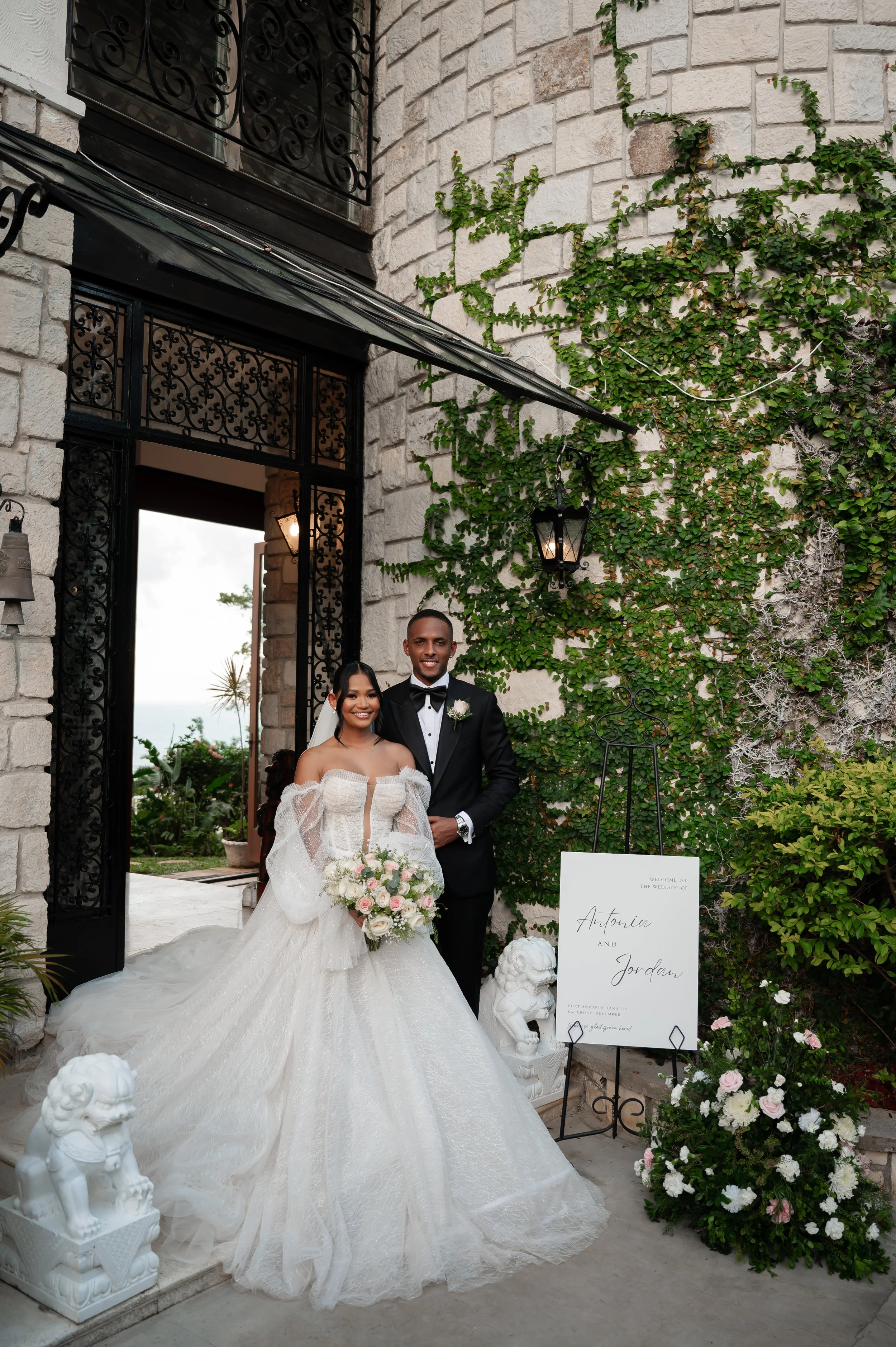 A newlywed couple standing in front of a wedding arch decorated with green ivy and flowers, with a sign that reads "Welcome to the wedding of Antonia and Jordan". The bride is wearing a strapless white wedding gown with off-the-shoulder sleeves, hold