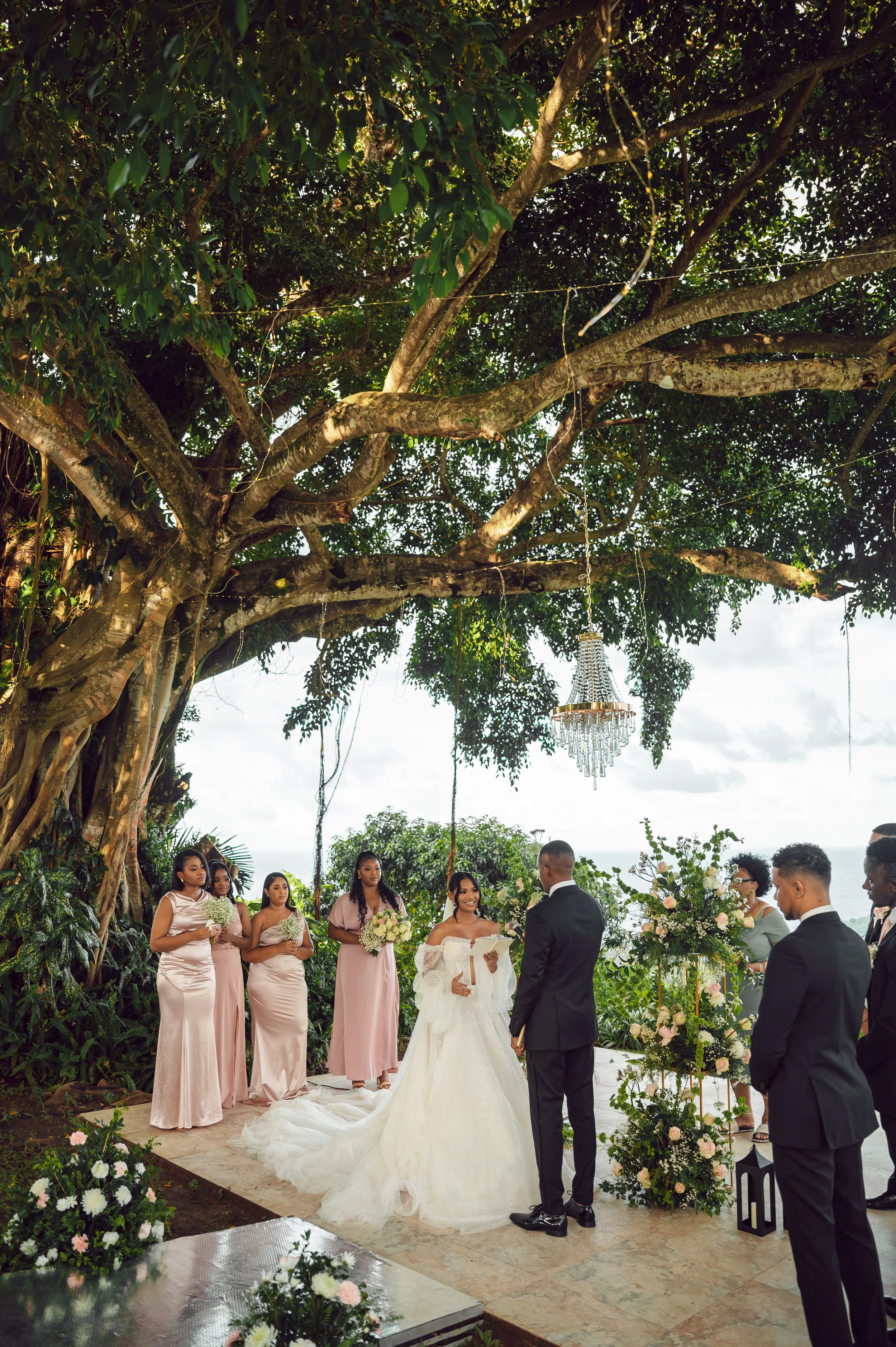 A wedding ceremony taking place outdoors under a large tree with green leaves, with the bride and groom facing each other and surrounded by bridesmaids and groomsmen. The ceremony is decorated with floral arrangements and hanging lights, with a sceni