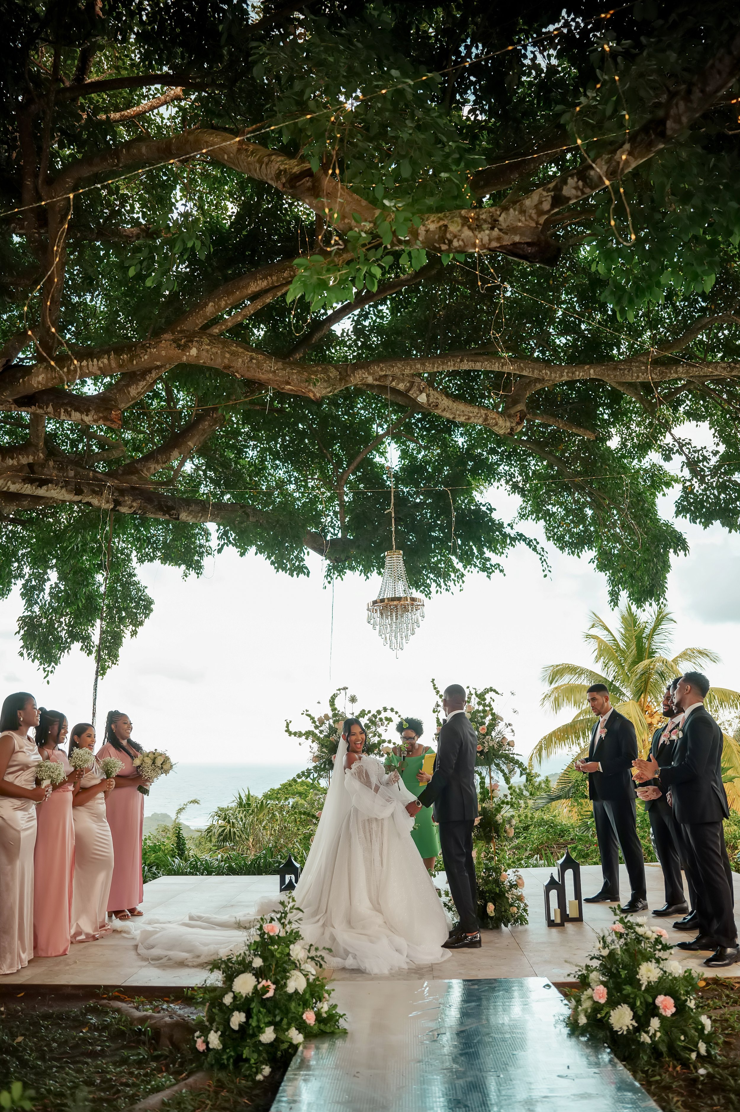 A wedding ceremony taking place outdoors under a large tree decorated with string lights and a chandelier. The bride and groom are holding hands at the altar, with bridesmaids on the left holding bouquets and wearing pink dresses, and groomsmen on th