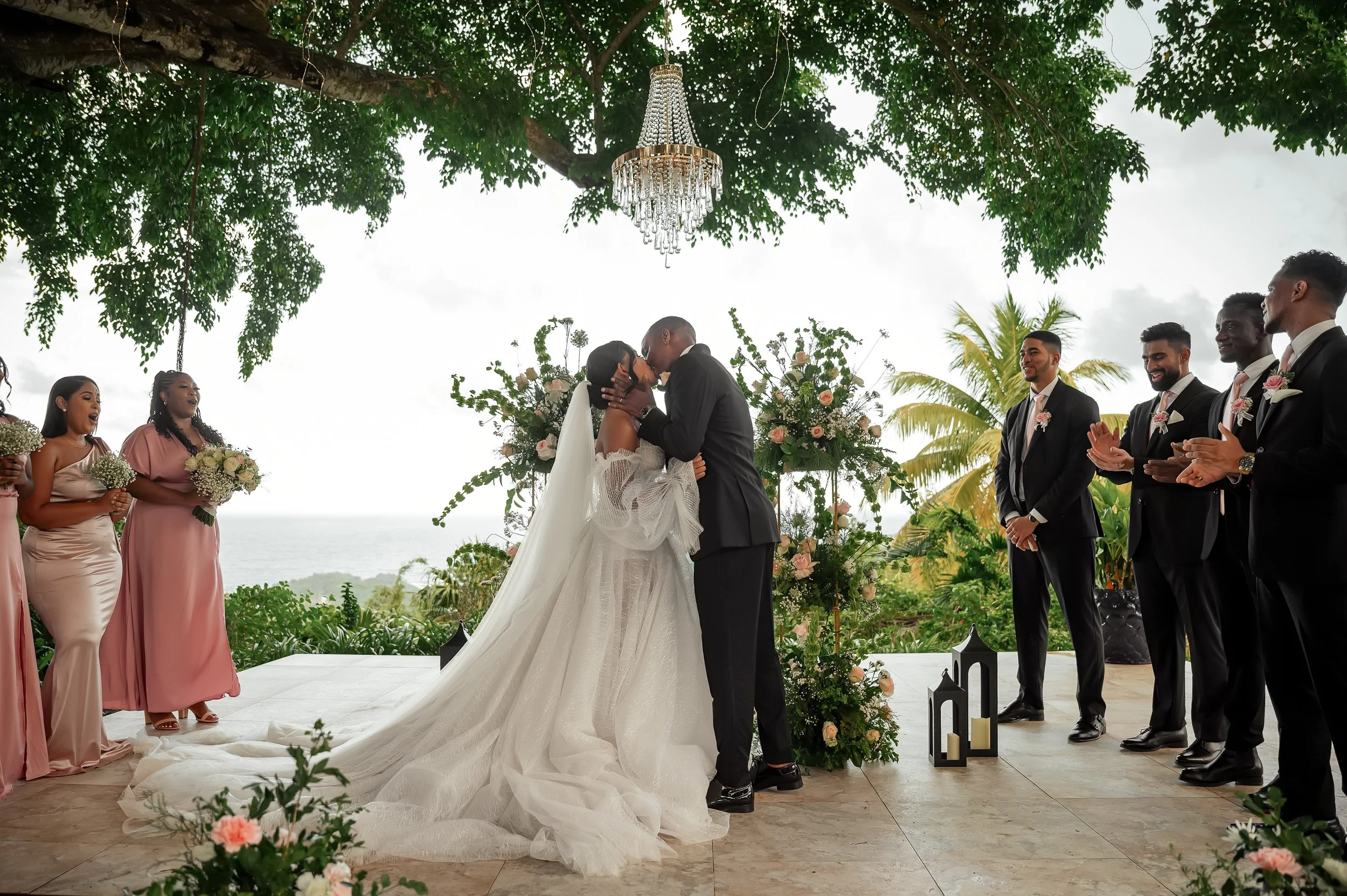 A wedding ceremony outdoors with a bride and groom kissing under a tree with a chandelier, surrounded by bridesmaids and groomsmen, with lush greenery and the ocean in the background.