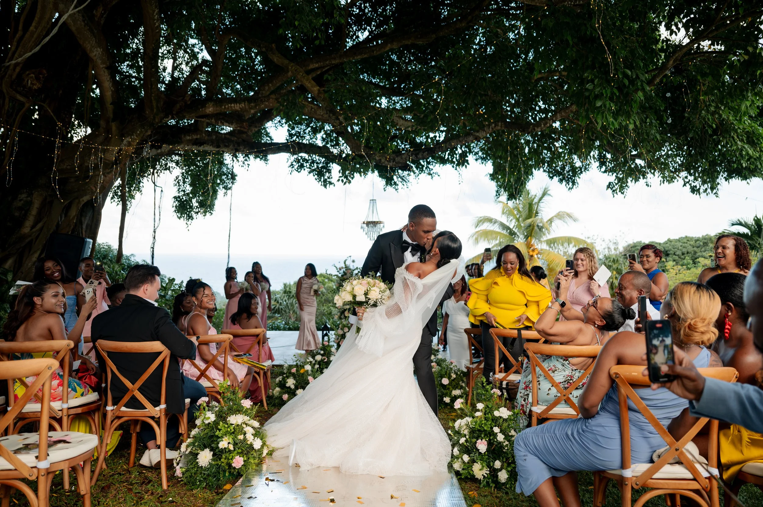 Bride and groom kissing during outdoor wedding ceremony under a large tree with string lights, surrounded by seated guests taking photos, with ocean view in the background.