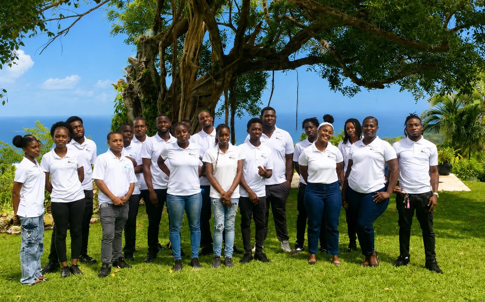 A group of approximately twenty people standing outside on grass in front of a large tree with hanging roots, with a view of the ocean and blue sky in the background. They are wearing white shirts and are smiling at the camera.