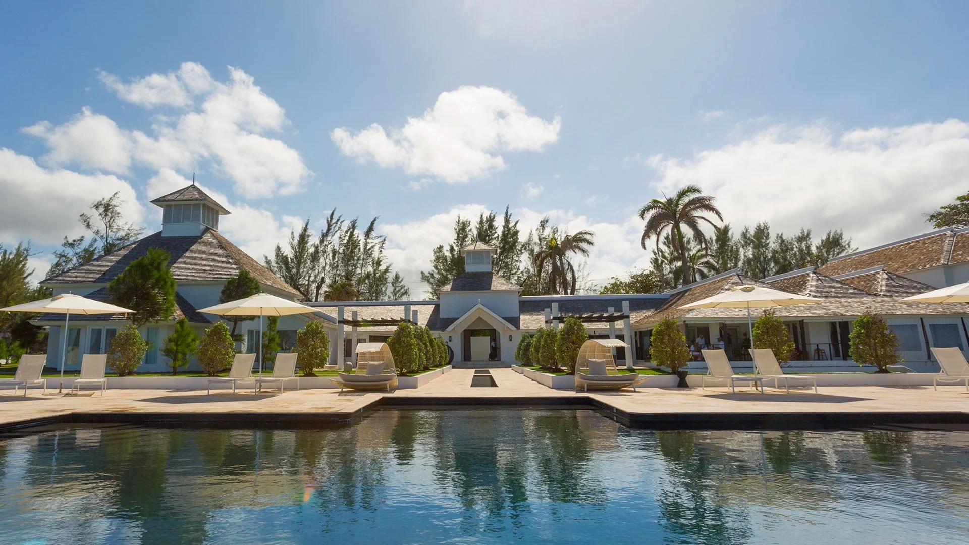 A luxurious resort with a large outdoor swimming pool in the foreground, surrounded by lounge chairs and umbrellas. The resort buildings have a modern design with white walls and shingle roofs. There are tall palm trees and lush greenery around the buildings, with a bright blue sky and fluffy clouds overhead.
